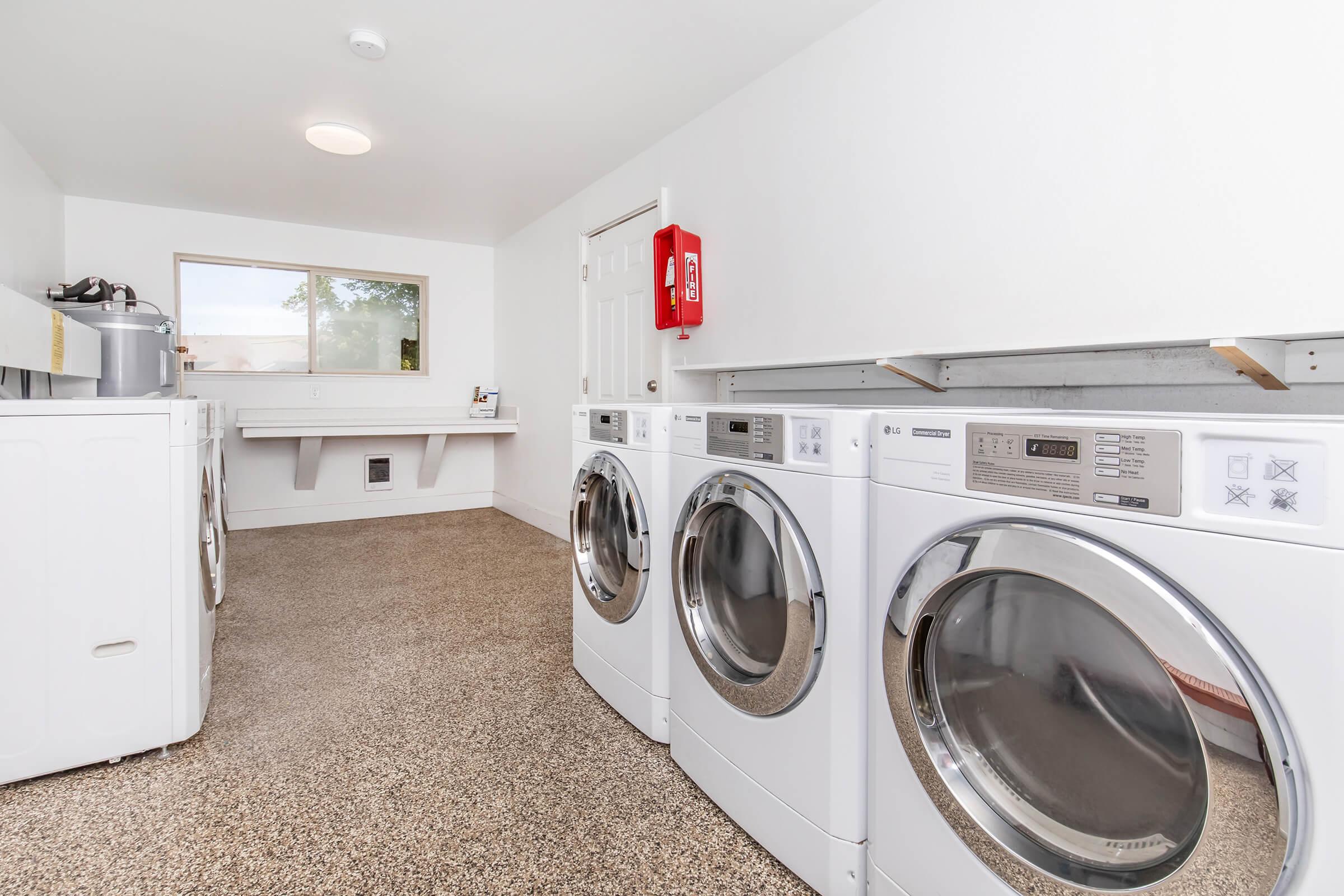 A clean laundry room featuring three front-loading washing machines and a dryer. There is a countertop for folding clothes, a window allowing natural light, and a fire extinguisher mounted on the wall. The floor is finished with a smooth surface.