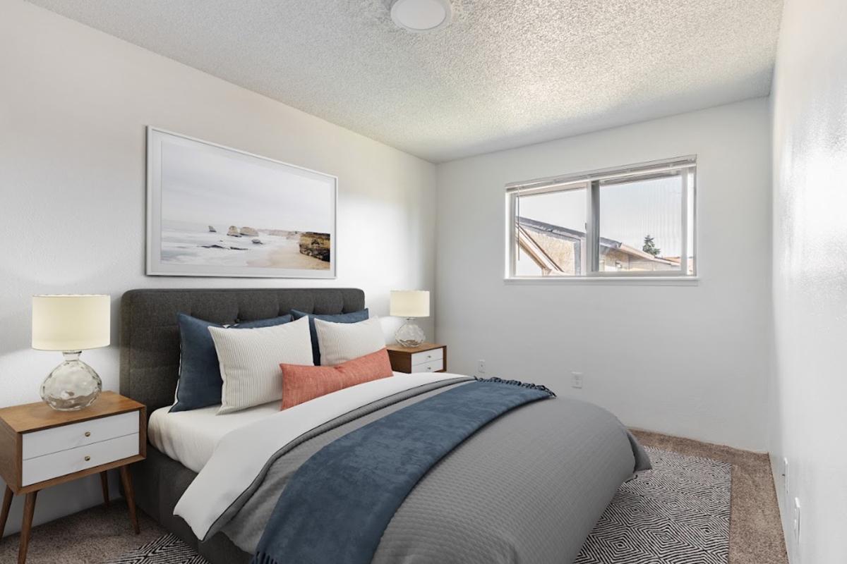 Cozy bedroom featuring a comfortable bed with gray and white bedding, decorative pillows in shades of blue and coral, two bedside tables with lamps, and a large window allowing natural light. A framed landscape picture hangs above the bed, enhancing the serene ambiance. Walls are painted white, and carpeted flooring adds warmth.