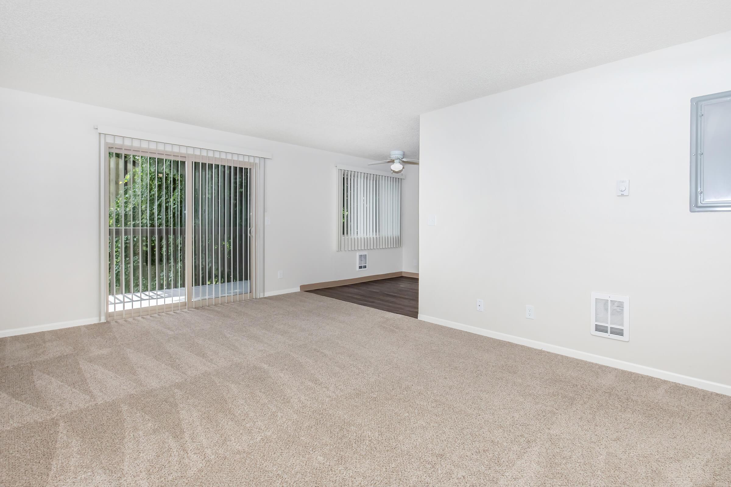 Spacious living area with light beige carpet, neutral-colored walls, and large sliding glass doors leading to an outdoor space. Window blinds are partially open, allowing natural light to brighten the room. A ceiling fan is visible, and a separate room is seen in the background.