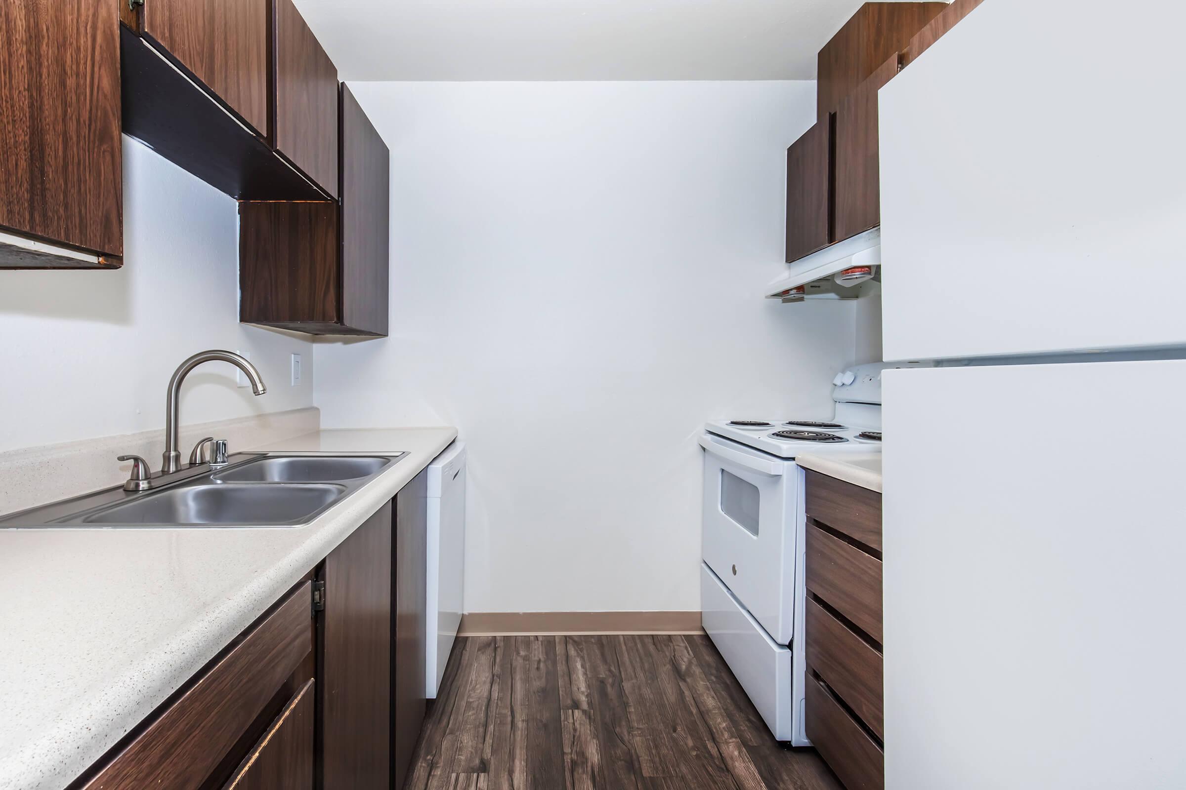 A modern kitchen featuring dark wooden cabinets, a double sink, a white refrigerator, and a white oven. The spacious layout includes a countertop with a glossy finish and minimalist design, complemented by a light-colored wall and dark wood flooring. The kitchen appears clean and well-maintained.