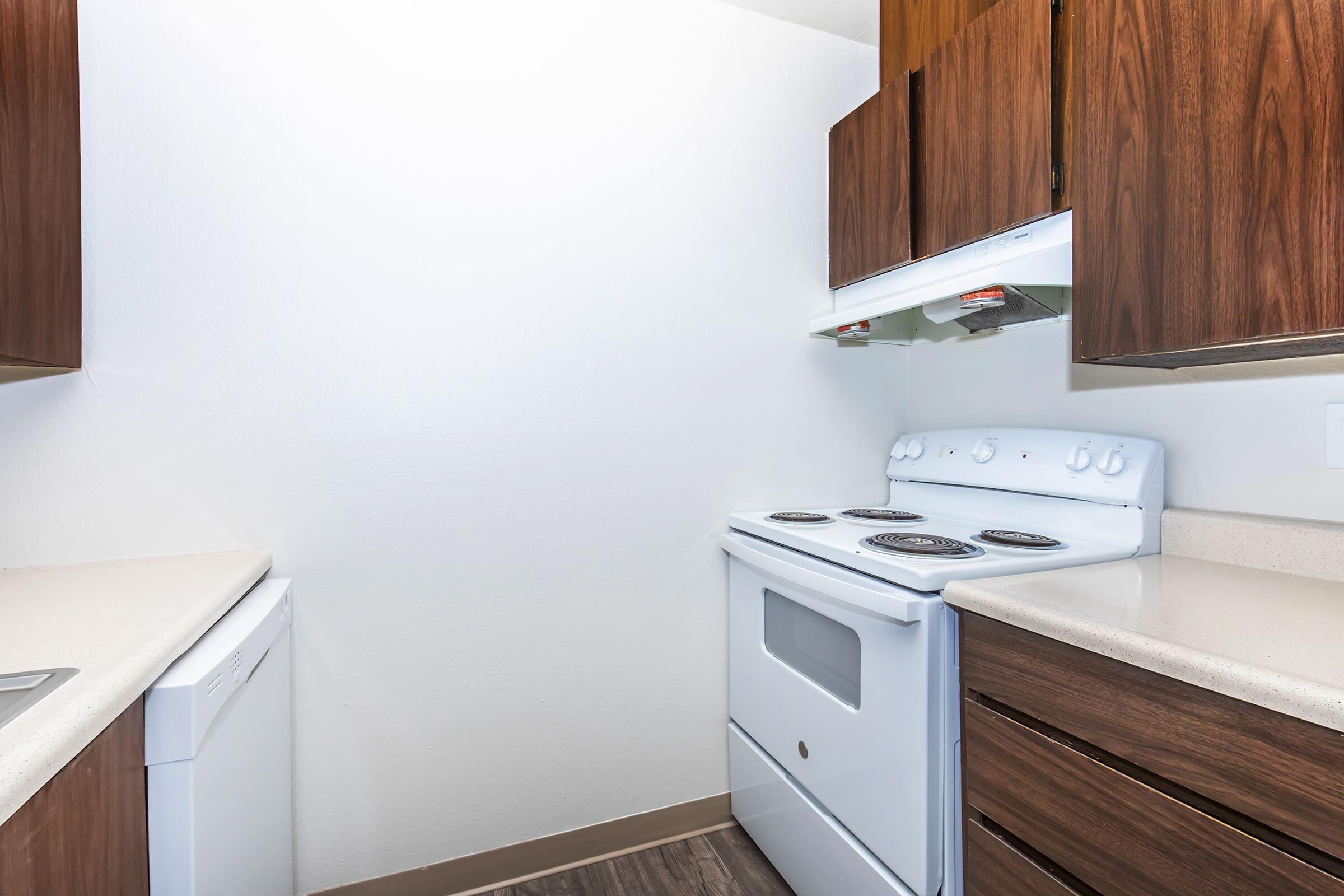 A modern kitchen featuring a white stove and oven, a hood vent, a dishwasher, and dark wooden cabinets. The countertops are light-colored and the walls are plain white. The layout suggests a compact space, with appliances and cabinetry arranged efficiently.