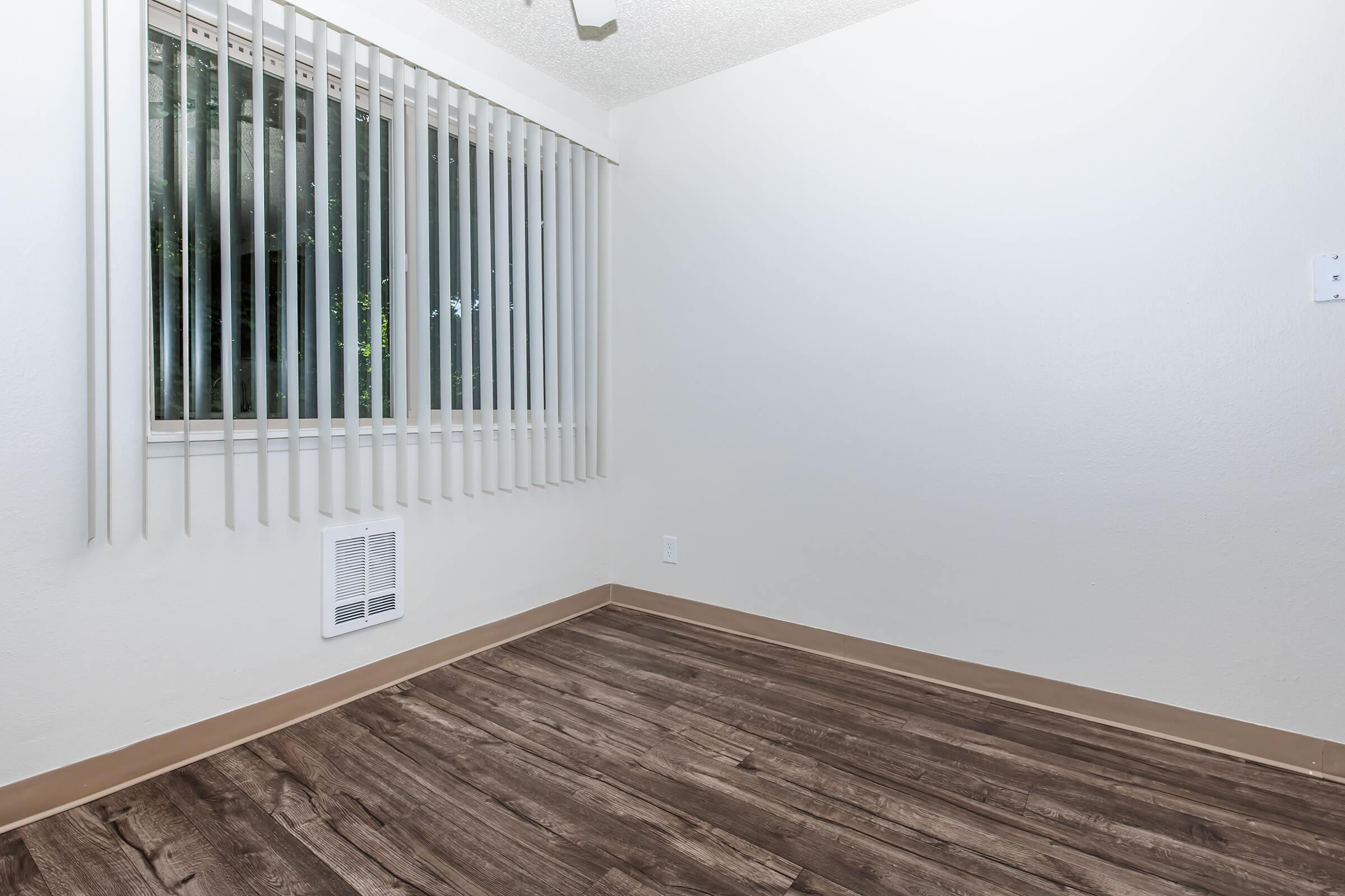 Empty room with light-colored walls and a window featuring vertical blinds. The floor is made of dark vinyl planks, and there is an air vent on one wall. The space appears bright and minimalistic, with no furniture present.