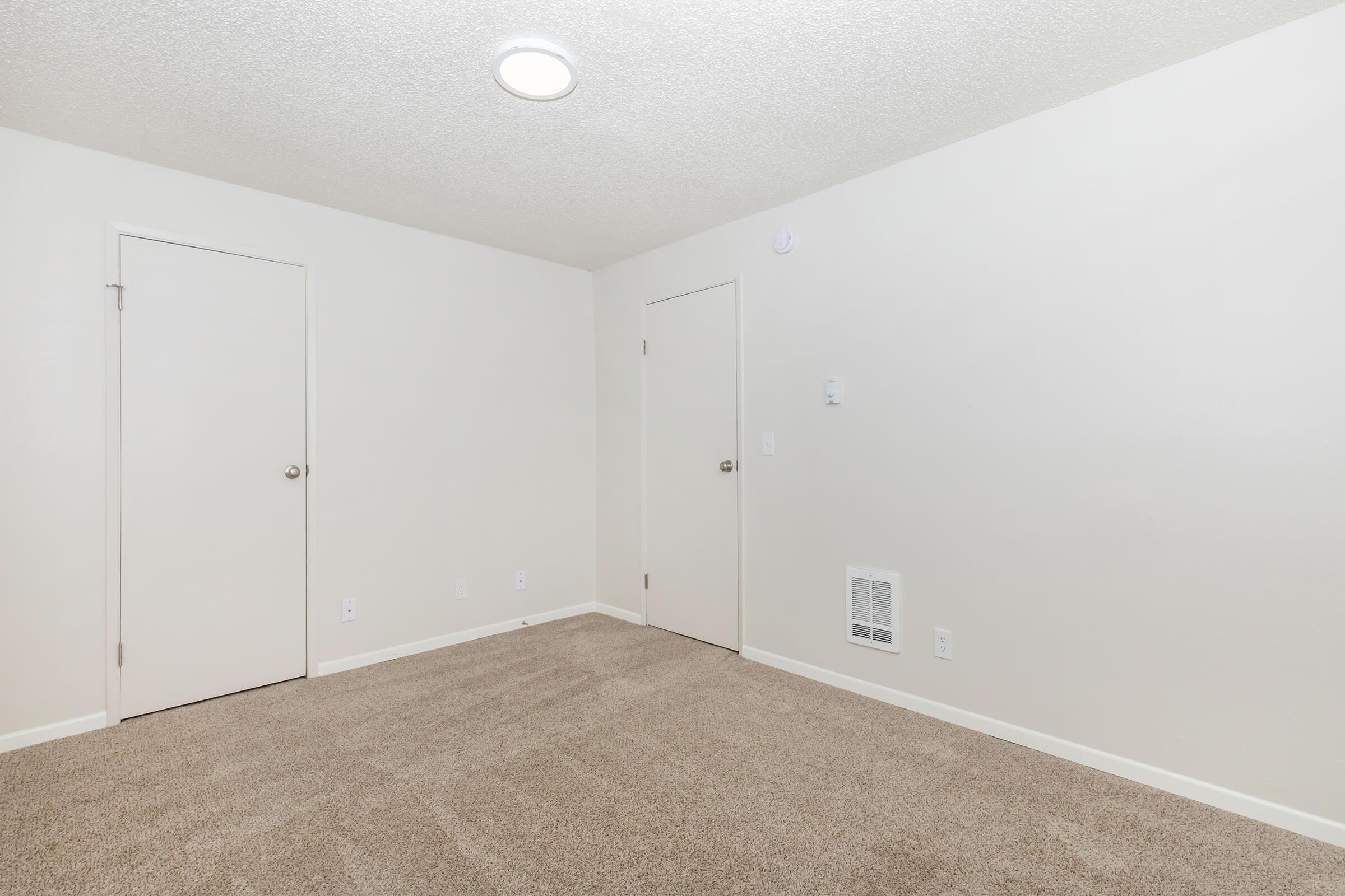 Empty, well-lit room with beige carpet and off-white walls. Two doors are visible on the left and right sides, and a ceiling light provides illumination. The space appears clean and minimalistic, suitable for various uses such as a bedroom or office.