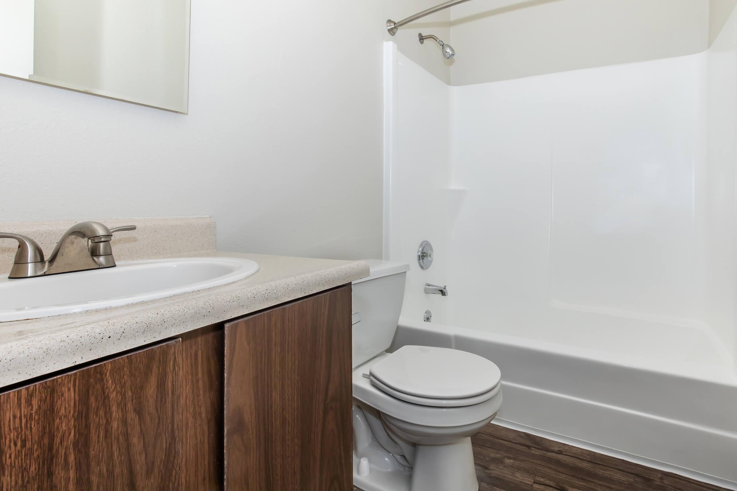 A clean and modern bathroom featuring a white bathtub and shower combo, a vanity with a sink, and wooden cabinetry. The walls are painted in a light color, and there is a mirror above the sink. The flooring is a wood-like laminate, enhancing the overall fresh and contemporary appearance of the space.