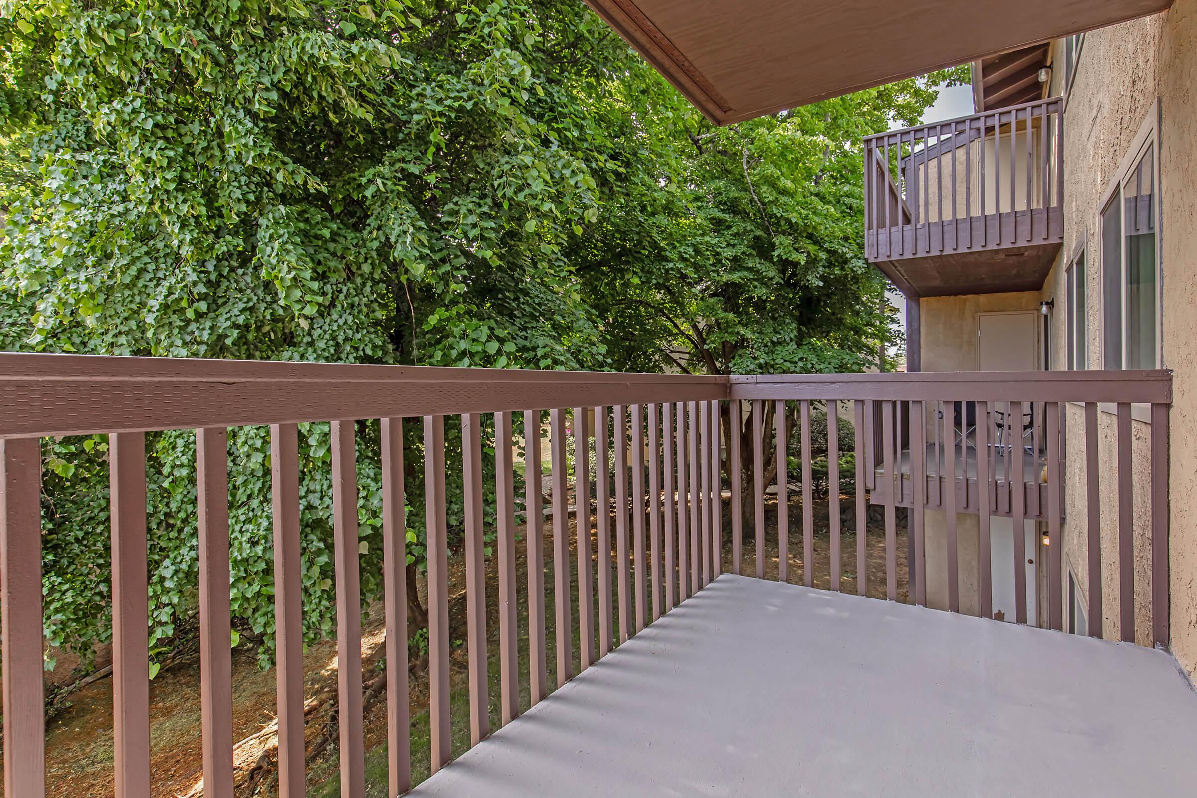 A view from a balcony showing a wooden railing and a spacious concrete floor. Lush green trees are visible in the background, providing a natural, shaded setting. Two additional balconies can be seen on adjacent buildings, creating a cozy and inviting atmosphere.