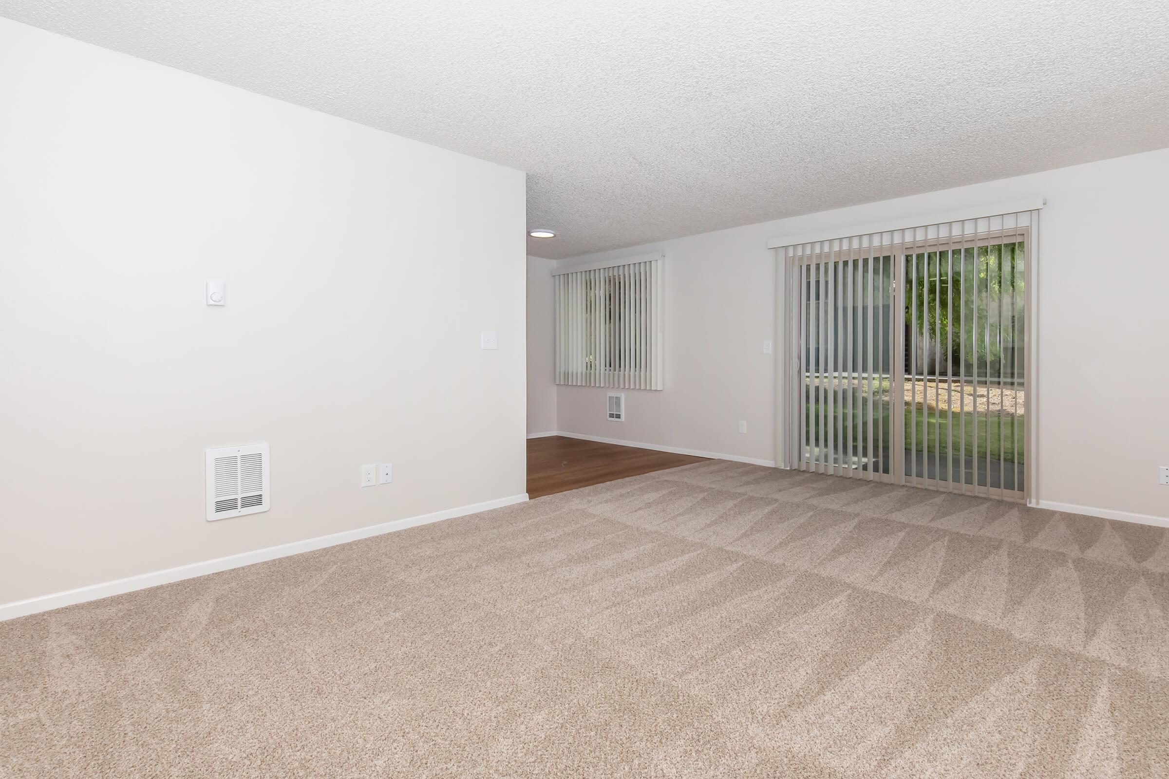 A spacious, empty room featuring beige carpet flooring and light gray walls. There are vertical blinds on a sliding glass door that leads to an outdoor area. The ceiling has a textured finish, and there is a heating vent on one wall. The overall ambiance is bright and inviting, perfect for customization.