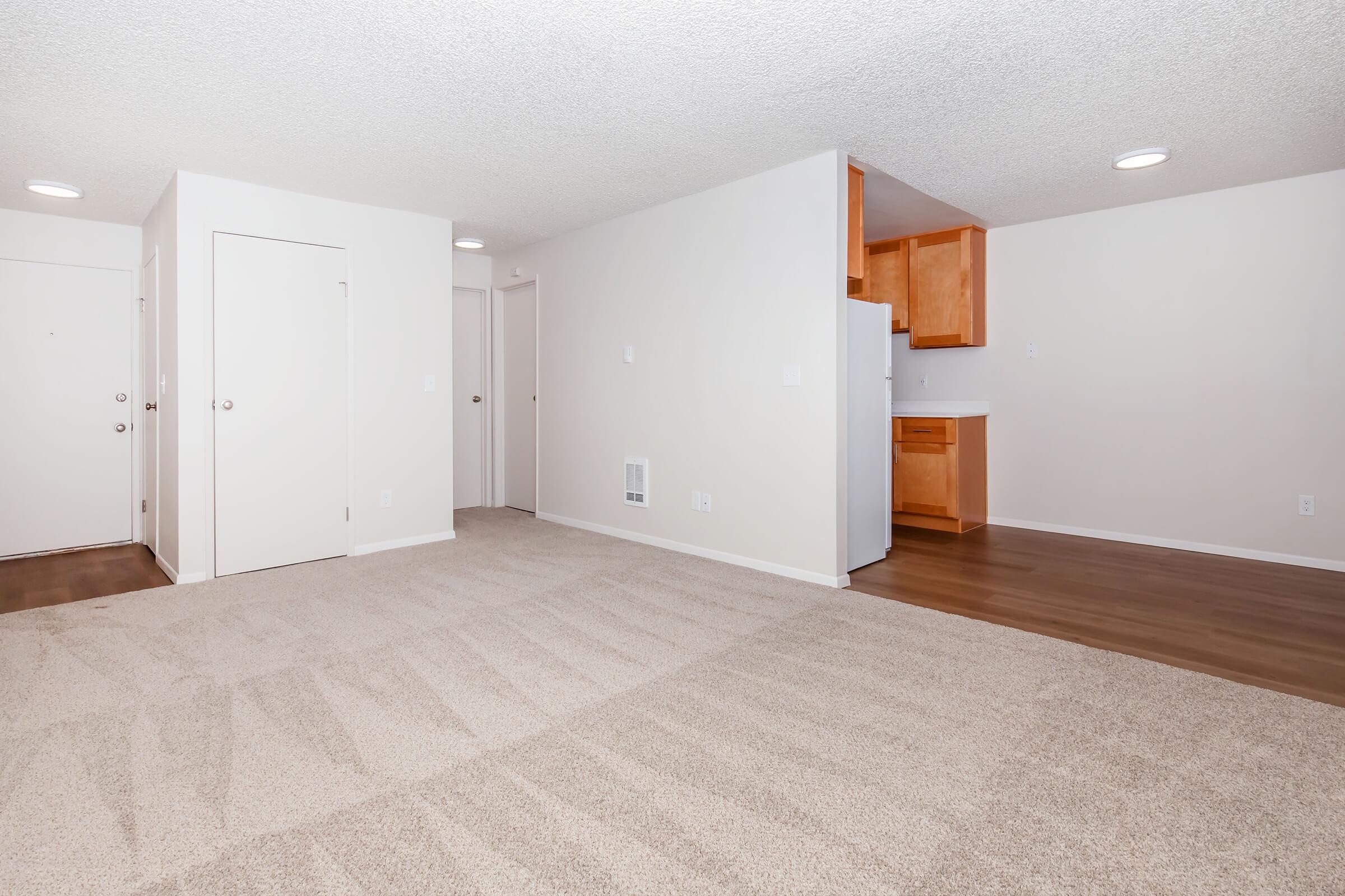 A spacious, empty living area with carpeted flooring leading to a kitchen. The walls are painted in a light color, and there are doors on the left side, one leading outside and the other likely to a closet. The kitchen in the background features wooden cabinetry and appliances, creating an open and airy atmosphere.