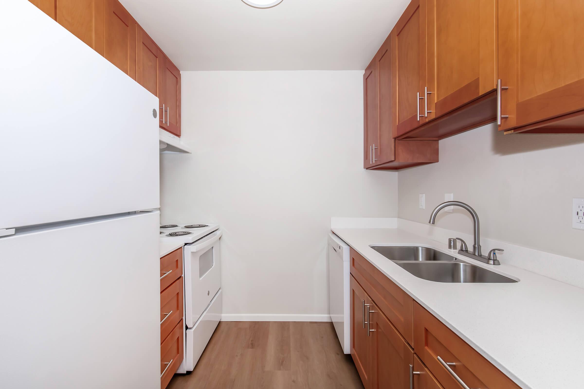 Modern kitchen featuring light wood cabinets, a white refrigerator, electric stove, and a double sink with a sleek faucet. The countertops are white, and the flooring is a light wood finish, creating a bright and spacious atmosphere. The walls are painted in a neutral shade.
