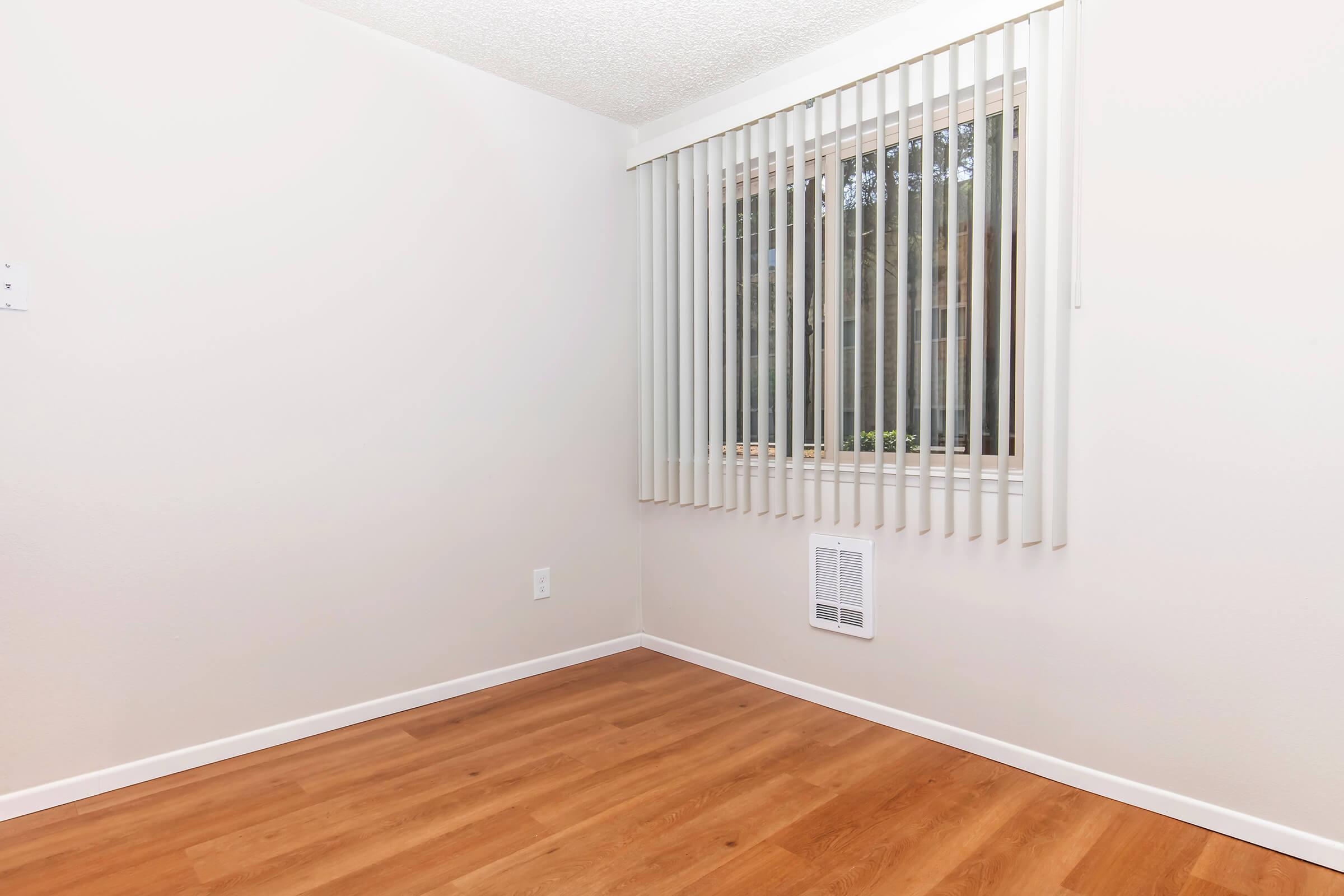 A bright, empty room with light-colored walls and wooden flooring. A window with vertical blinds is positioned on one wall, allowing natural light to enter. A small air vent is visible near the floor, and the overall atmosphere is clean and minimalistic.