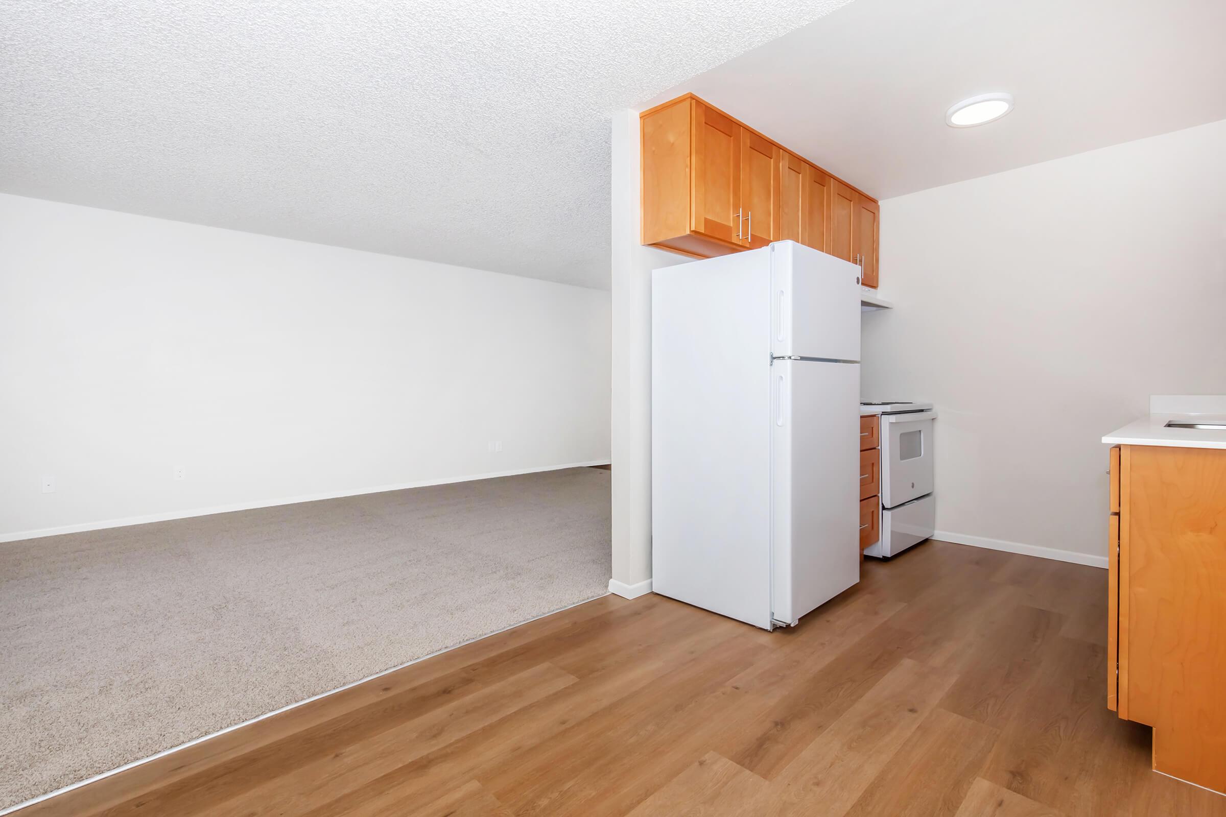 A partially furnished kitchen area featuring a white refrigerator, an oven, and light wood cabinetry. The adjacent space is carpeted and mostly empty, with a plain white wall and ceiling. The flooring is a warm wooden tone that contrasts with the light fixtures above.