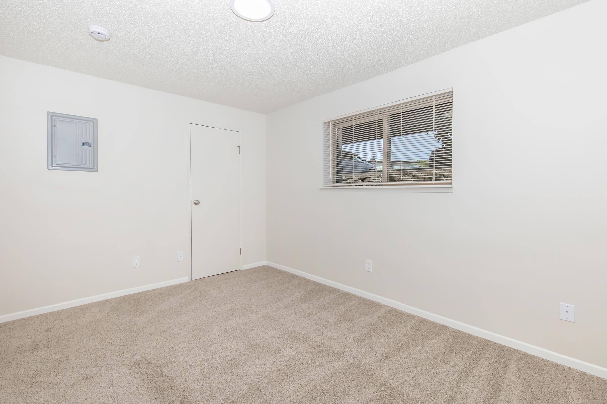 Empty room with beige carpet, light-colored walls, and a window with blinds. A white door is present on one wall, and an electrical panel is mounted near the ceiling on the opposite wall. The room is well-lit with natural light coming through the window.