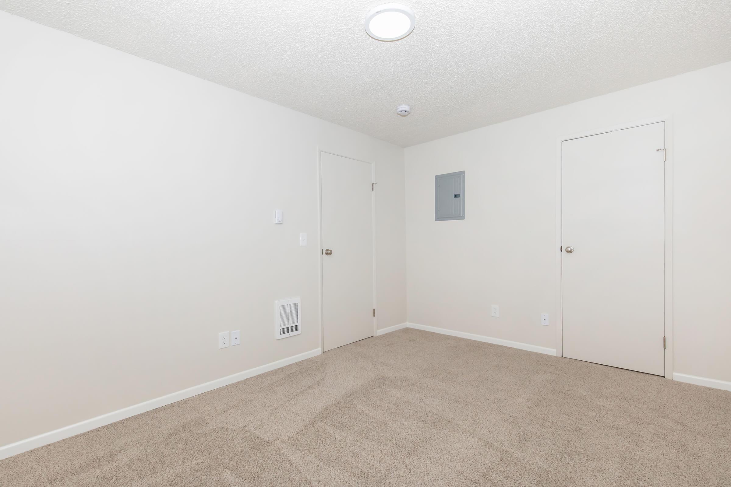 Empty room with light-colored walls and beige carpet. There are two closed doors, one leading to a closet and the other possibly to another room. A wall-mounted electrical panel is visible, and the ceiling has a round light fixture. The space is uncluttered and minimalistic.