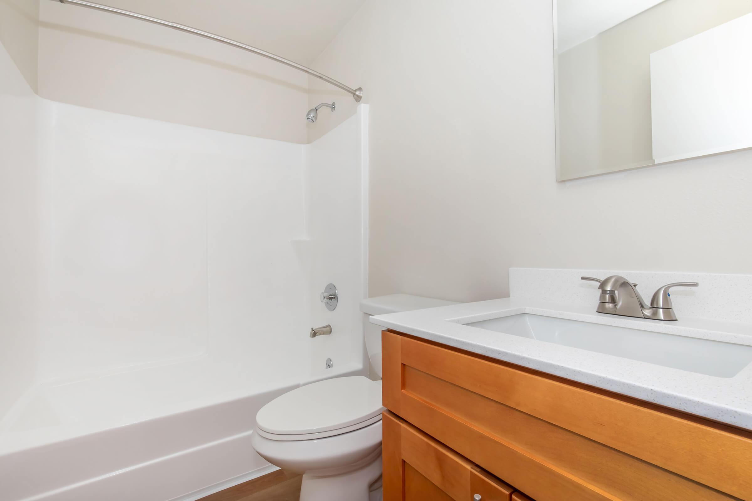 A clean and modern bathroom featuring a white shower stall, a toilet, and a wooden vanity with a white countertop and sink. A mirror is mounted on the wall above the sink. The walls are painted in a light color, creating a bright and airy atmosphere.