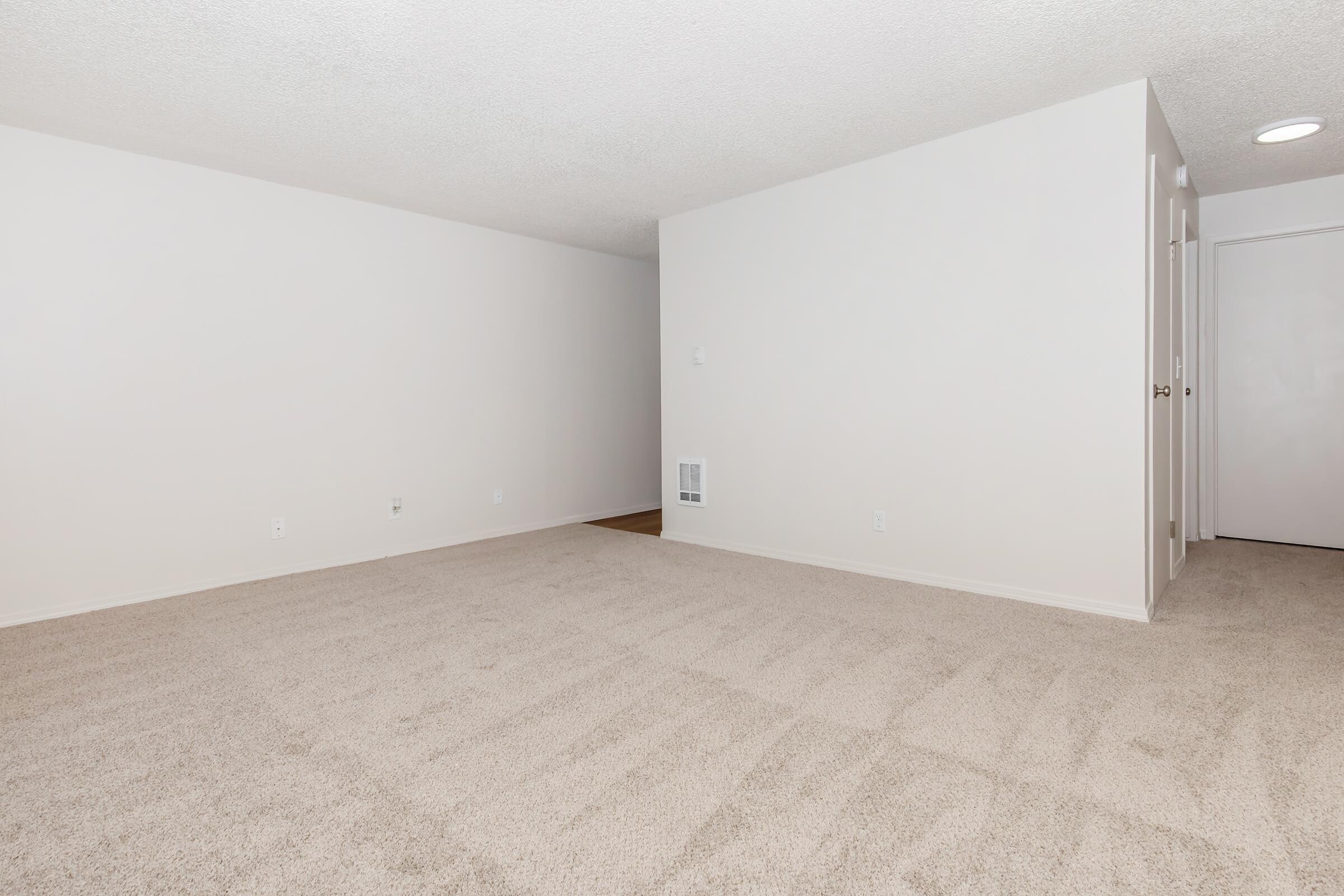 Empty room with light beige carpet and plain white walls. There is a small wall vent and a doorway on the right leading to another space. The lighting is soft, creating a bright and open atmosphere. The room is unadorned and lacks furniture, providing a blank canvas for decoration.