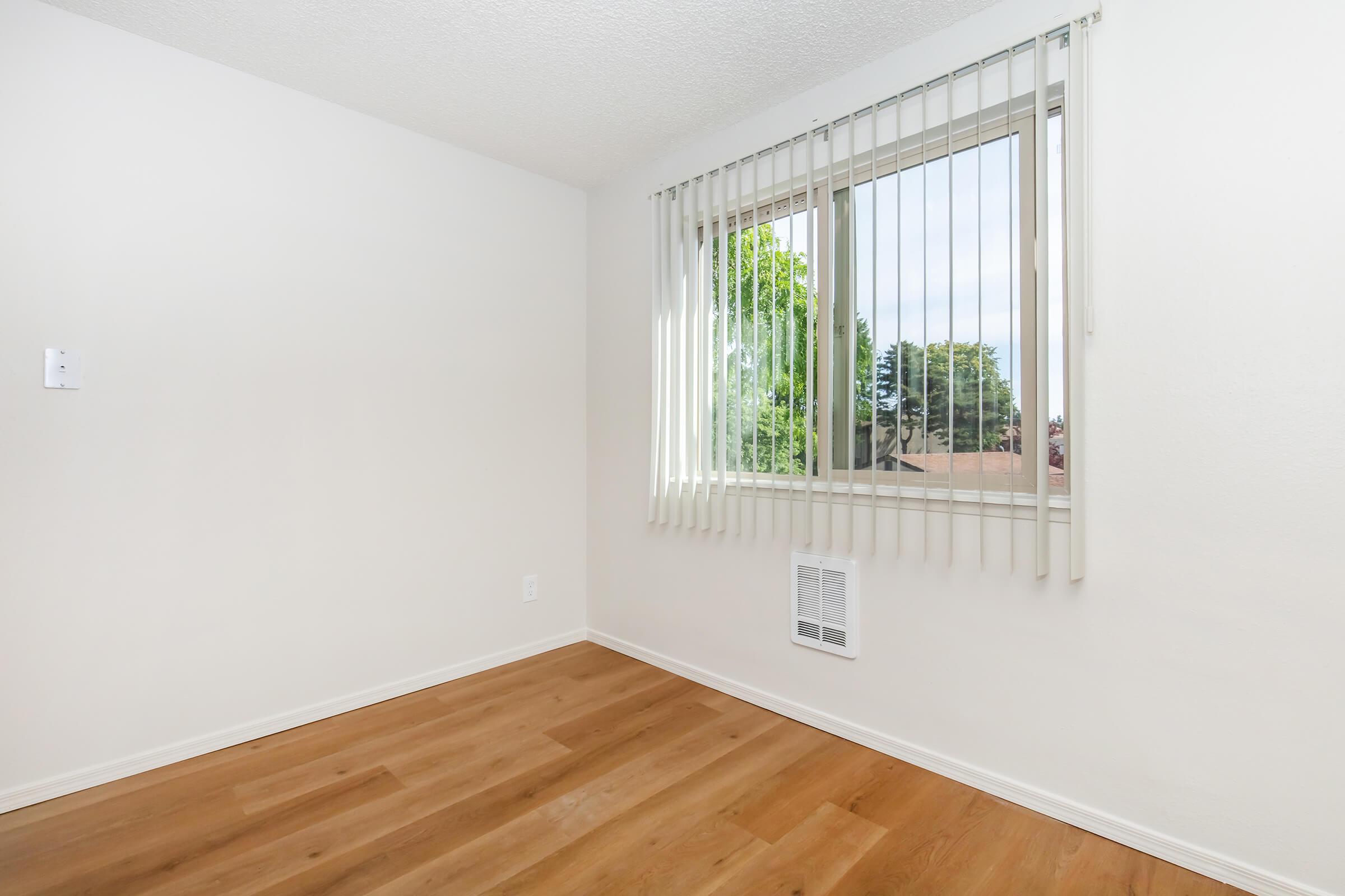 A bright, empty room with white walls and wooden flooring. It features a large window covered with vertical blinds, allowing natural light to enter. There's a wall-mounted heater beneath the window, and the space has a clean, minimalist appearance.