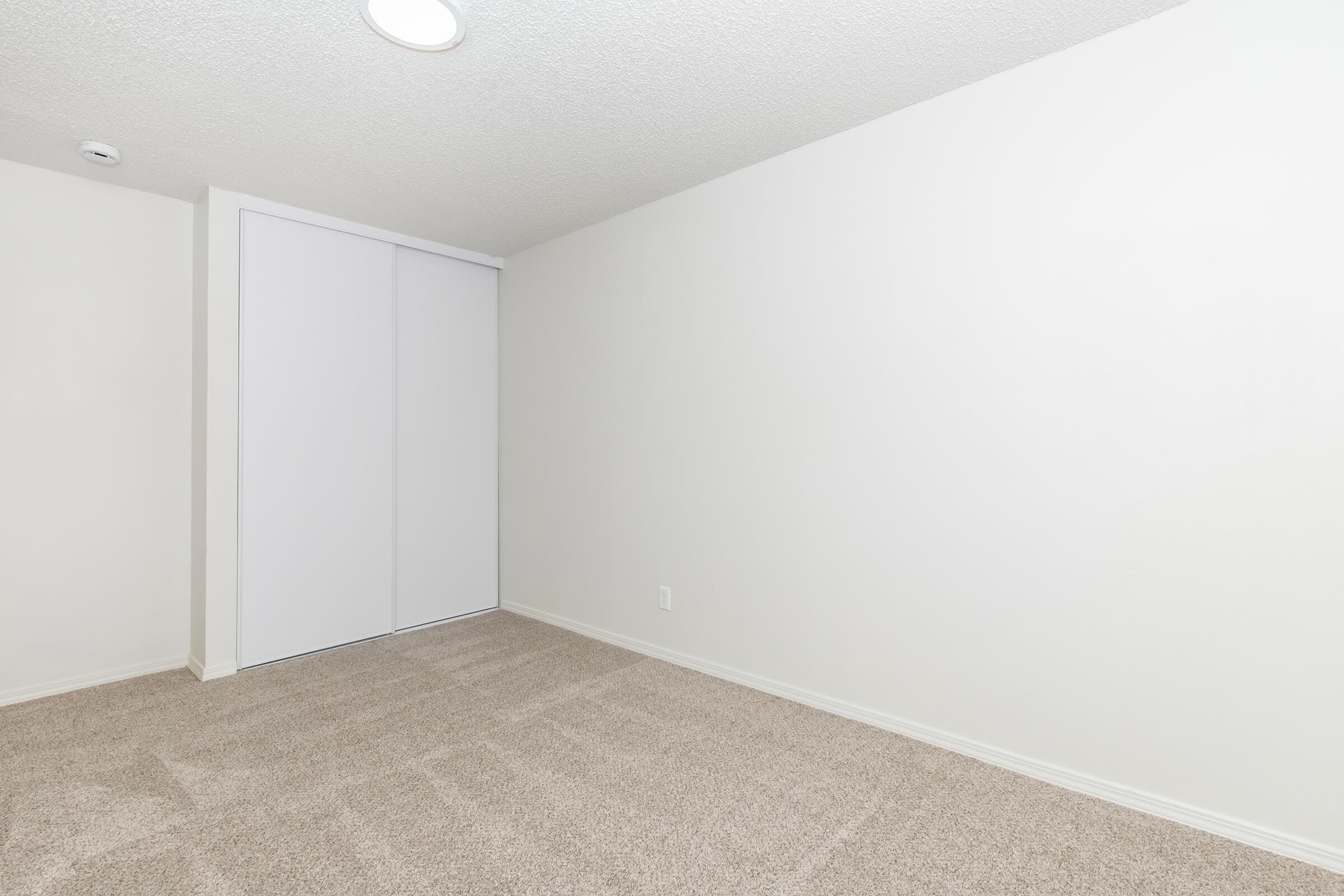 A bright, empty room with a textured ceiling and light-colored walls. The floor is covered in beige carpet, and there are sliding white closet doors on one side. The space is clean and minimalistic, ideal for various uses.