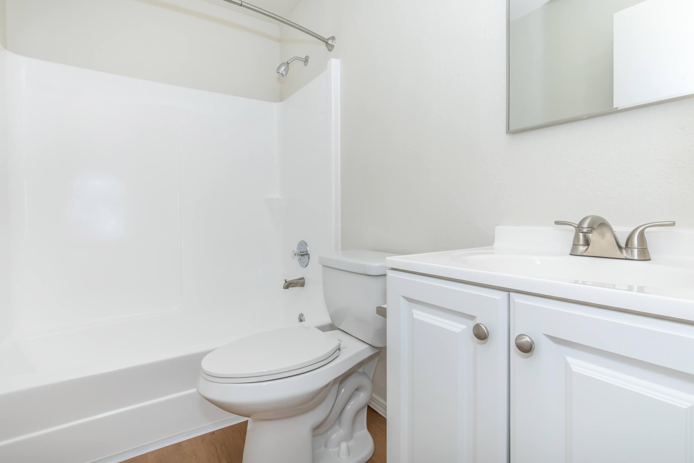 A clean and bright bathroom featuring a white bathtub with a shower, a white toilet, and a vanity with a sink. The walls are light-colored, and there is a mirror above the sink. The space is well-lit and has a simple, modern design.