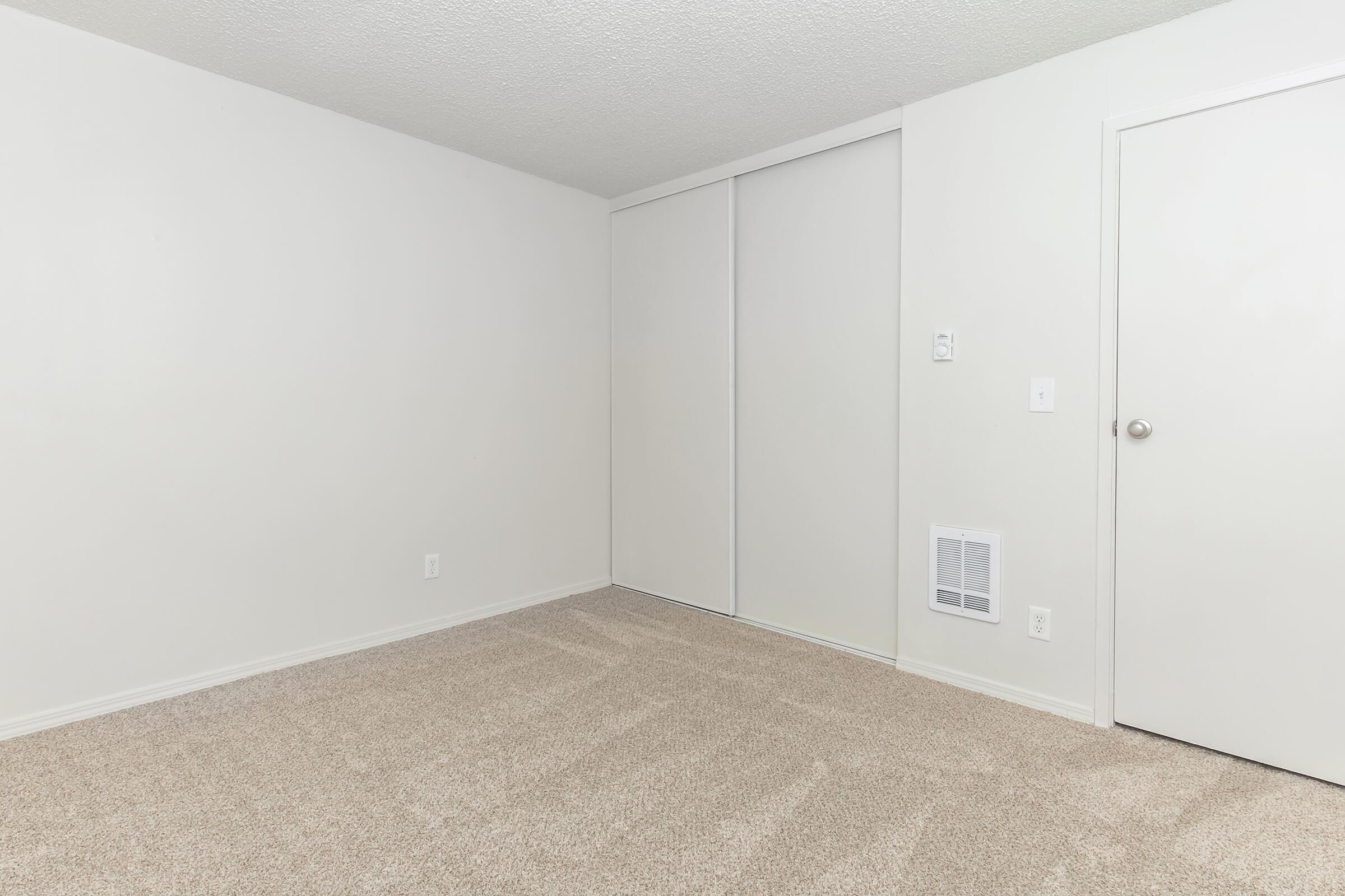 Empty room with light-colored walls and beige carpet. There is a closed white door on the right and a sliding closet door on the left. The room is well-lit, emphasizing its spaciousness and cleanliness.