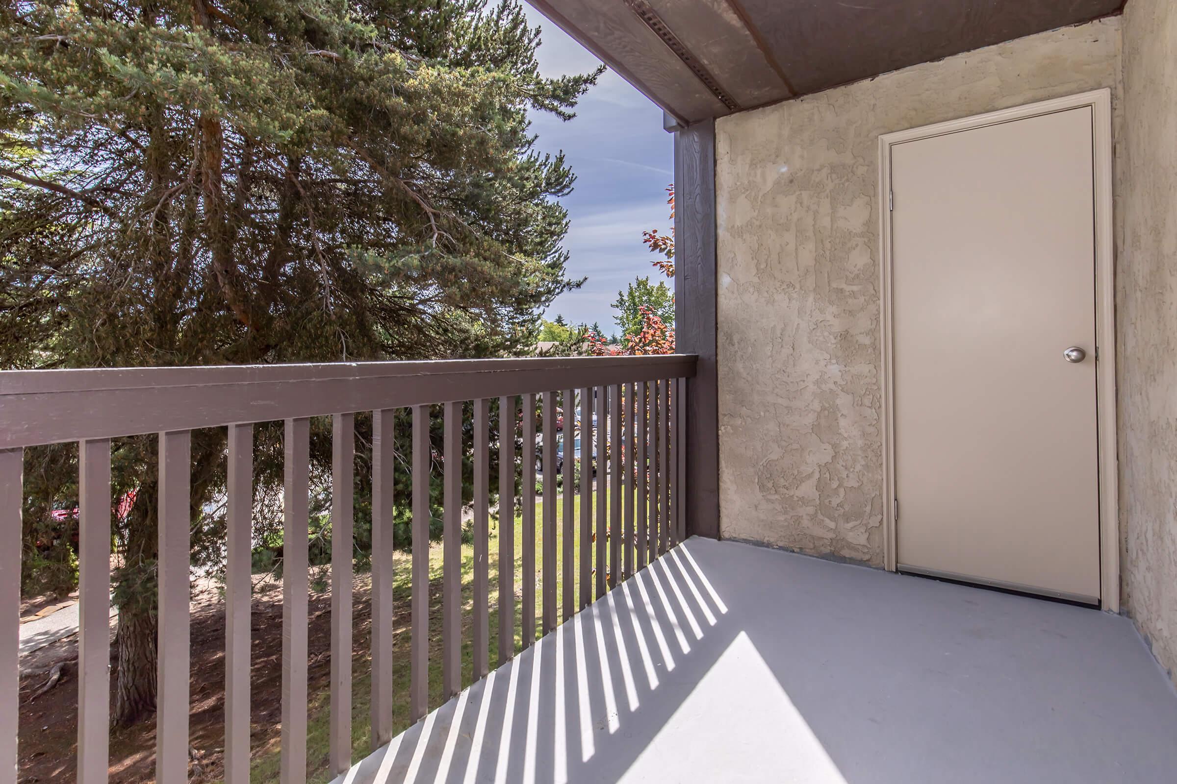 A balcony view featuring a wooden railing and a closed door. Sunlight casts shadows on the gray floor. In the background, there are green trees and foliage, suggesting a natural setting. The sky appears cloudy, indicating a possibly overcast day.