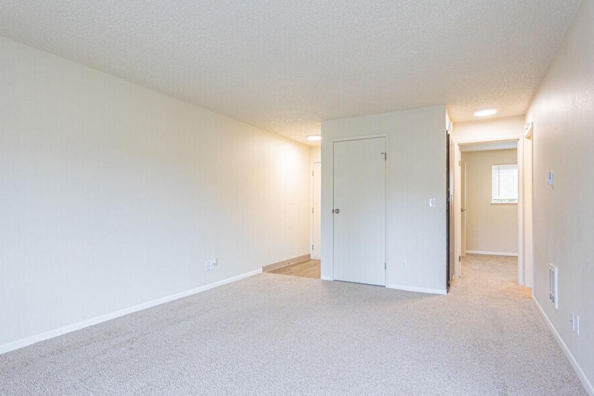 A spacious, empty room with beige walls and light gray carpeting. There’s a doorway leading to another area in the background, and a closet with a door to the right. Natural light enters from a window at the far end, creating a bright and airy atmosphere in the space.