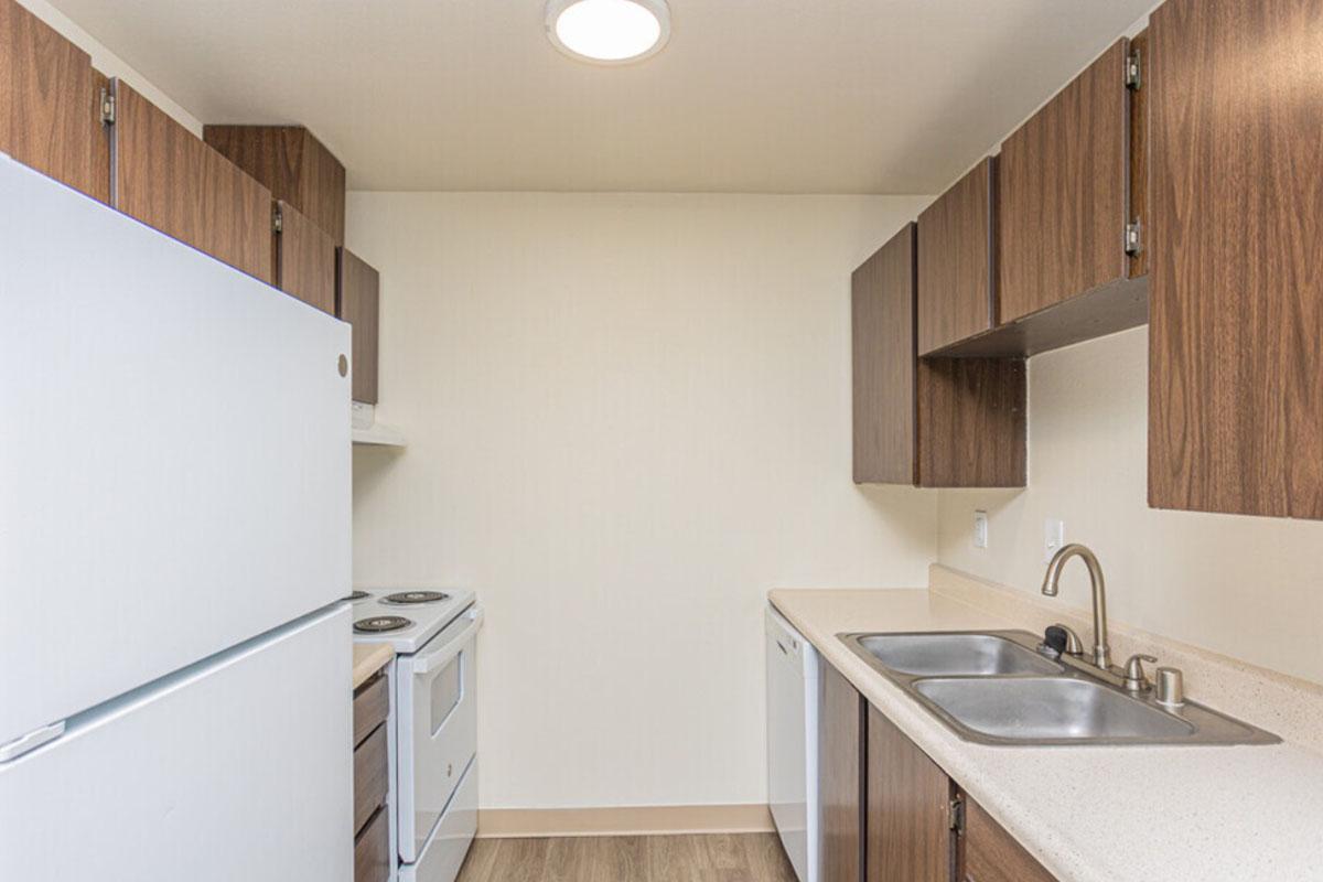 A small kitchen featuring a white refrigerator, a white stove, brown wooden cabinets, a light-colored countertop, and a double sink. The walls are plain and the floor has laminate wood flooring. The overall design is simple and functional, ideal for compact living spaces.