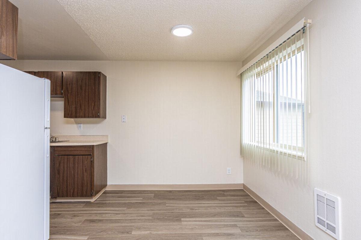 A bright, empty kitchen space featuring a white refrigerator, wooden cabinets, and a countertop. The walls are a light color and there are vertical blinds on a window, allowing natural light to fill the room. The floor is wood-patterned, creating a modern, clean aesthetic.