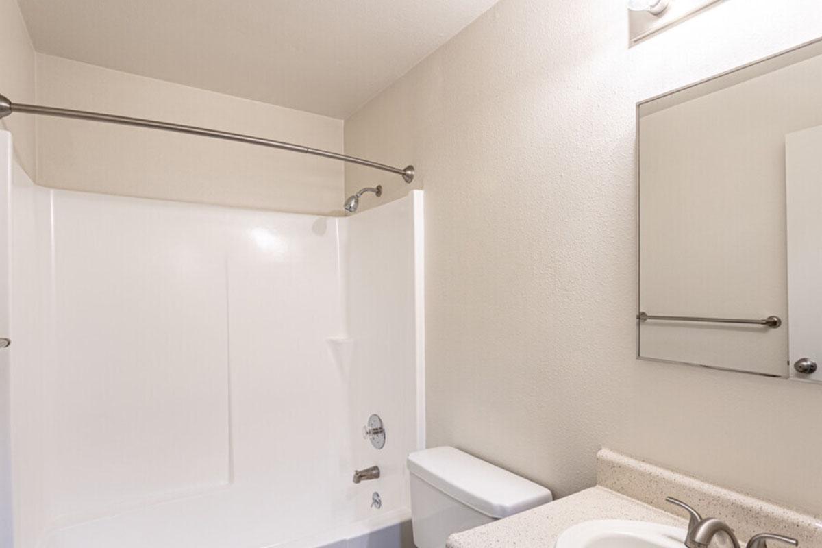 A clean, modern bathroom featuring a white bathtub with shower, a toilet, a countertop with a sink, and a large mirror. The walls are painted a neutral color, and there is a towel bar installed above the sink. Soft lighting enhances the overall brightness of the space.