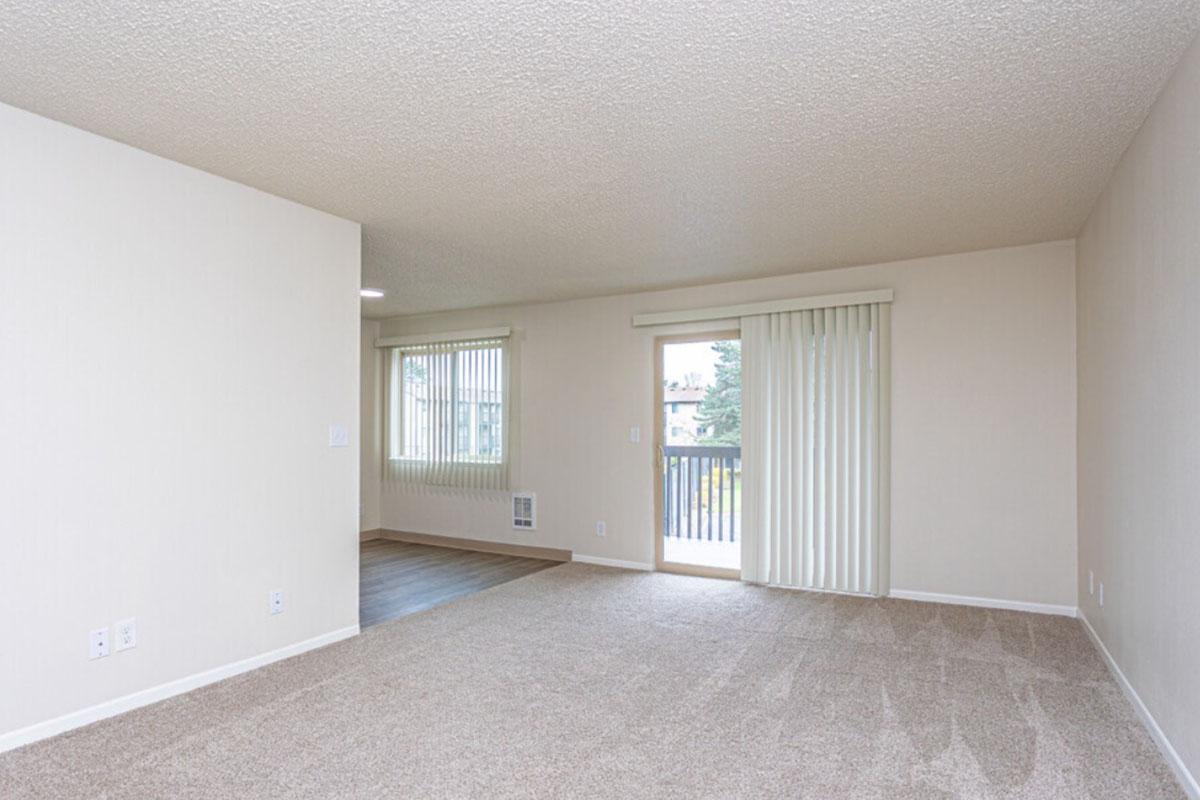 A spacious, unfurnished living area featuring light beige walls and carpet, a large window with vertical blinds letting in natural light, and a sliding glass door leading to a balcony. The room has an open layout, with another wall partially visible on the left.
