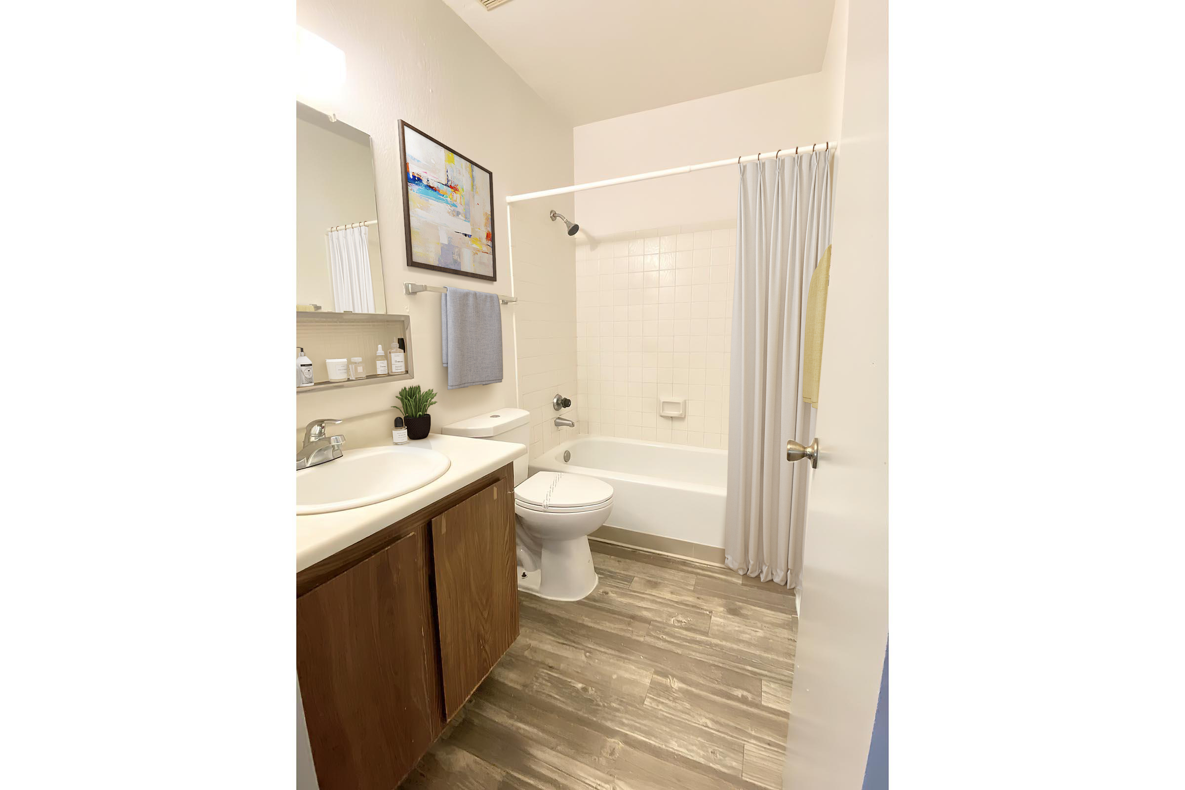 A clean and modern bathroom featuring a white bathtub and shower, a brown vanity with a sink, a mirror above the sink, and a small plant on the countertop. White shower curtain, light-colored tiles, and a neatly organized shelf with toiletries are visible. Natural light fills the space.