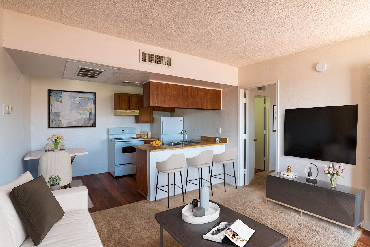 Cozy living space featuring a white sofa, coffee table, and a TV on a console. Open kitchen with wooden cabinets and bar seating for three. Natural light streams in through a window, enhancing the warm, inviting atmosphere. A touch of decor adds character to the room.