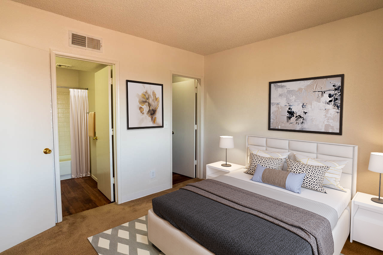 A cozy bedroom featuring a white bed with decorative pillows, a light gray blanket, and two bedside lamps. The room has neutral walls and a framed artwork. Doors lead to a bathroom and another room, with a shower curtain visible. A soft area rug adds warmth to the hardwood floor.