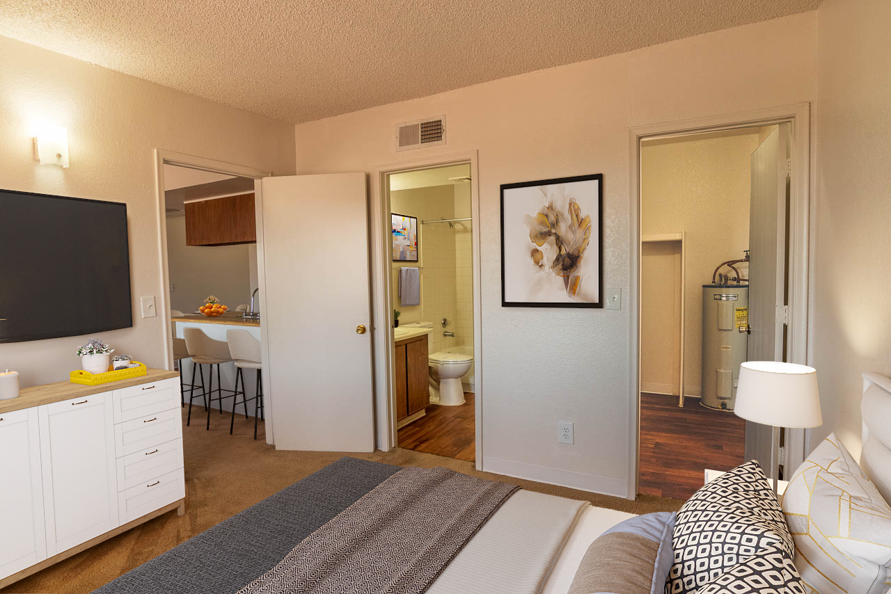 A cozy bedroom featuring a bed with decorative pillows, a small dresser with a TV, and a large piece of artwork on the wall. An open door leads to a bathroom, while another door opens to a hallway with a glimpse of a utility area. The walls are painted light, creating a warm ambiance.