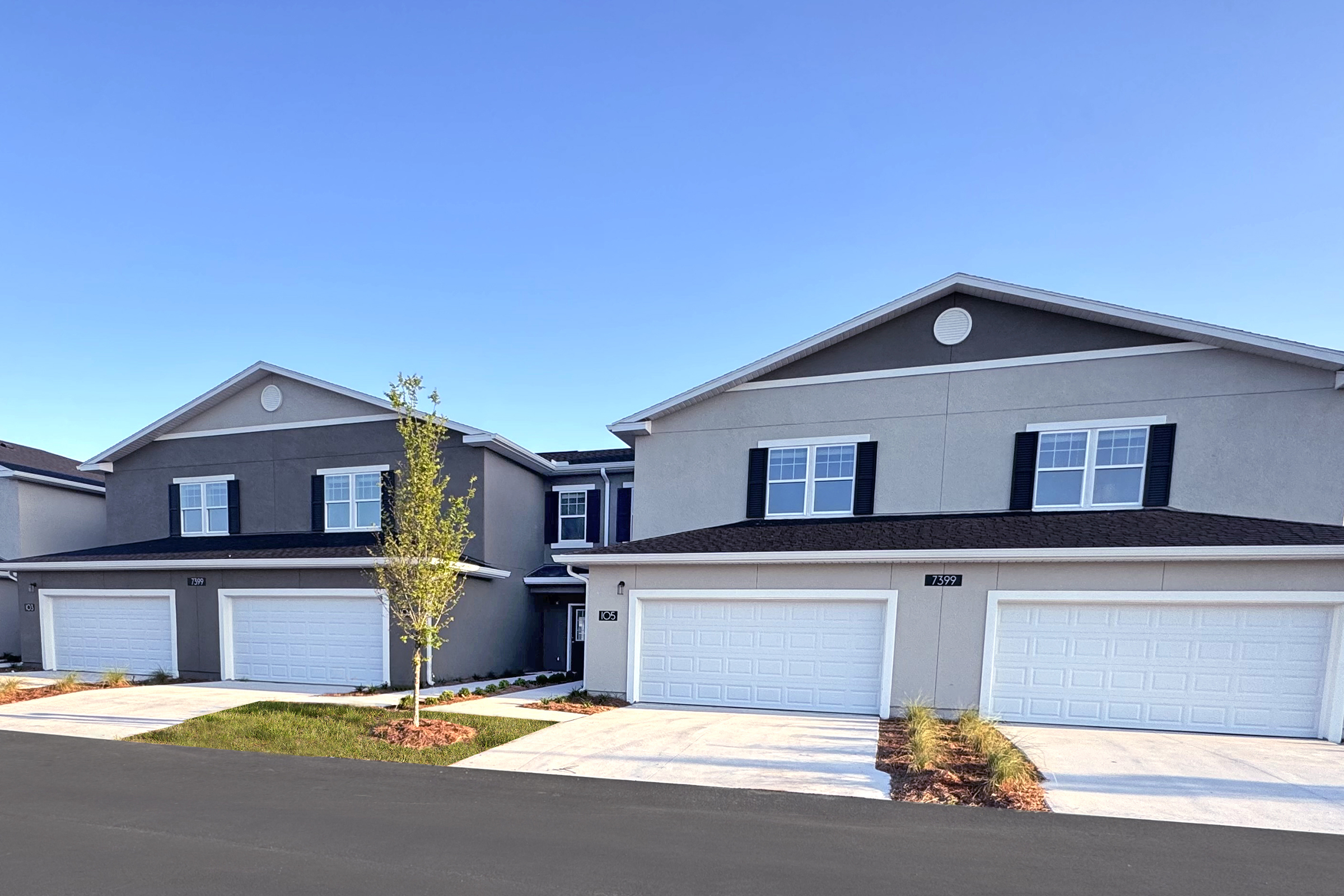 Newly constructed duplexes with modern architecture. Each unit features a double garage, symmetrical windows, and a neatly landscaped front yard. The sky is clear blue, complementing the light grey and dark grey facade of the buildings. The driveway is paved, leading to well-maintained grass and bushes.