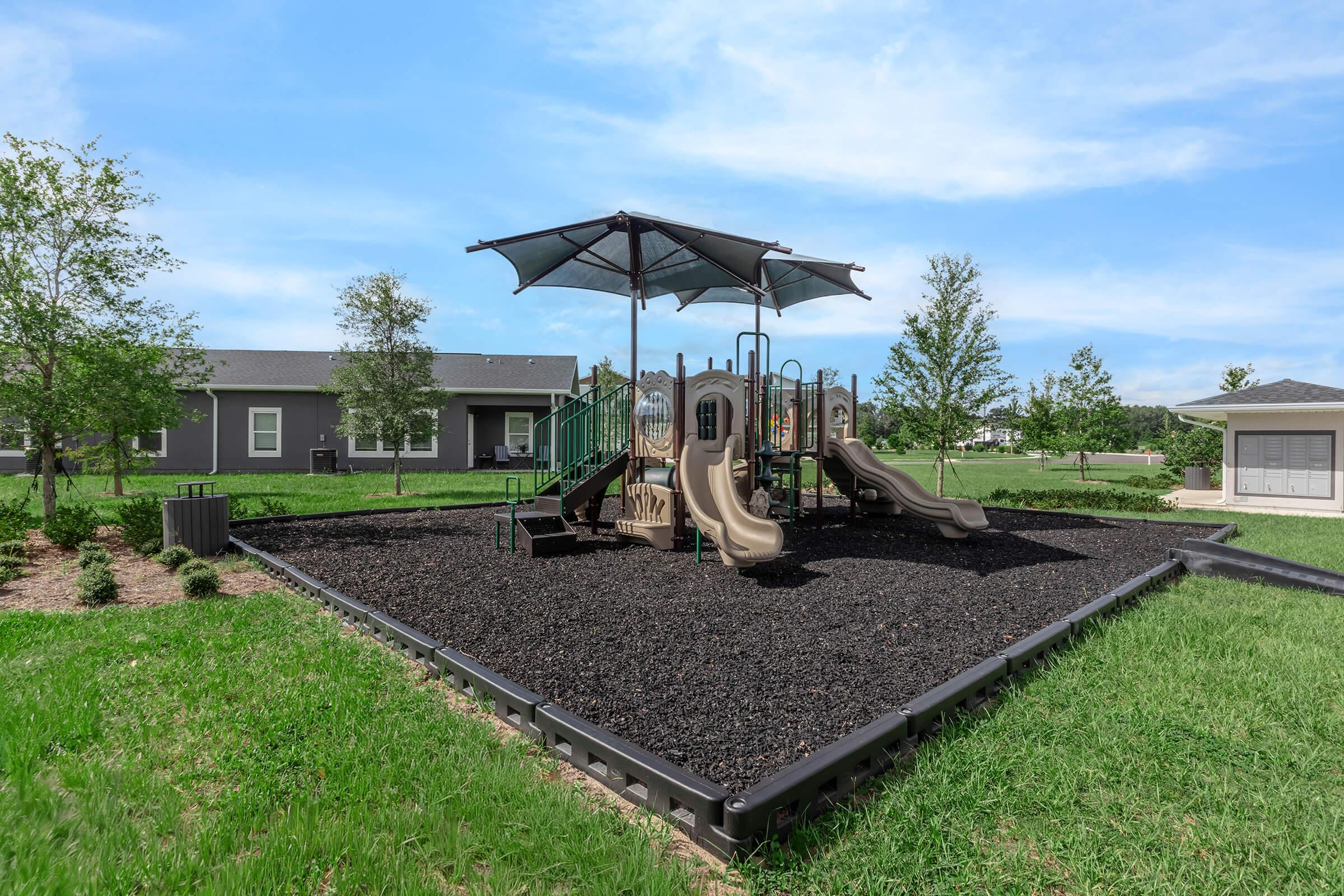 A playground featuring two slides, a climbing structure, and a large shaded canopy, surrounded by soft black mulch. The area is green with grass and small trees, set against a backdrop of residential buildings under a clear blue sky.