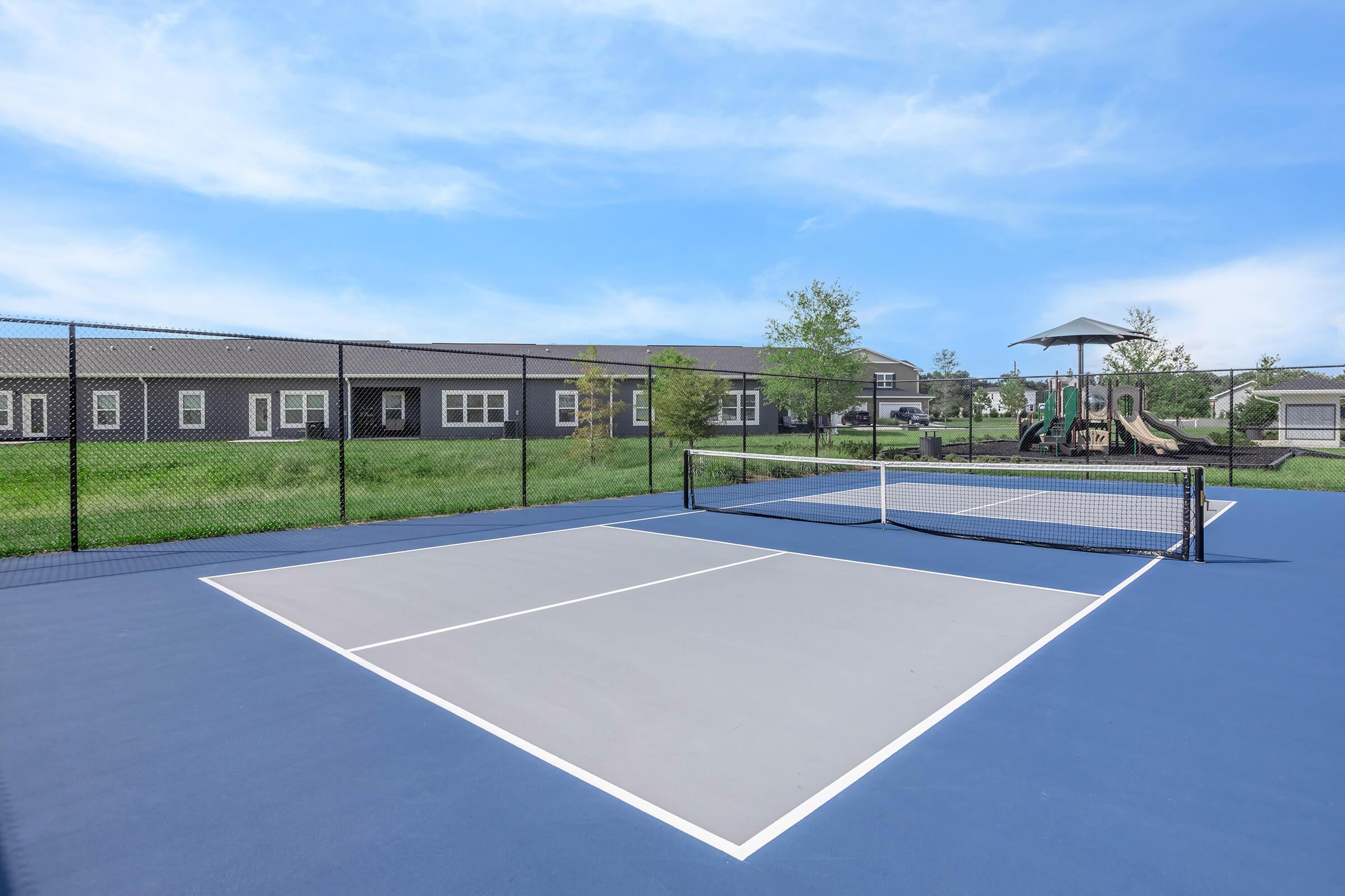 A tennis court with a blue surface and white lines, featuring a net in the center. In the background, there are several buildings and a playground with a slide, surrounded by grass and trees, under a clear blue sky.