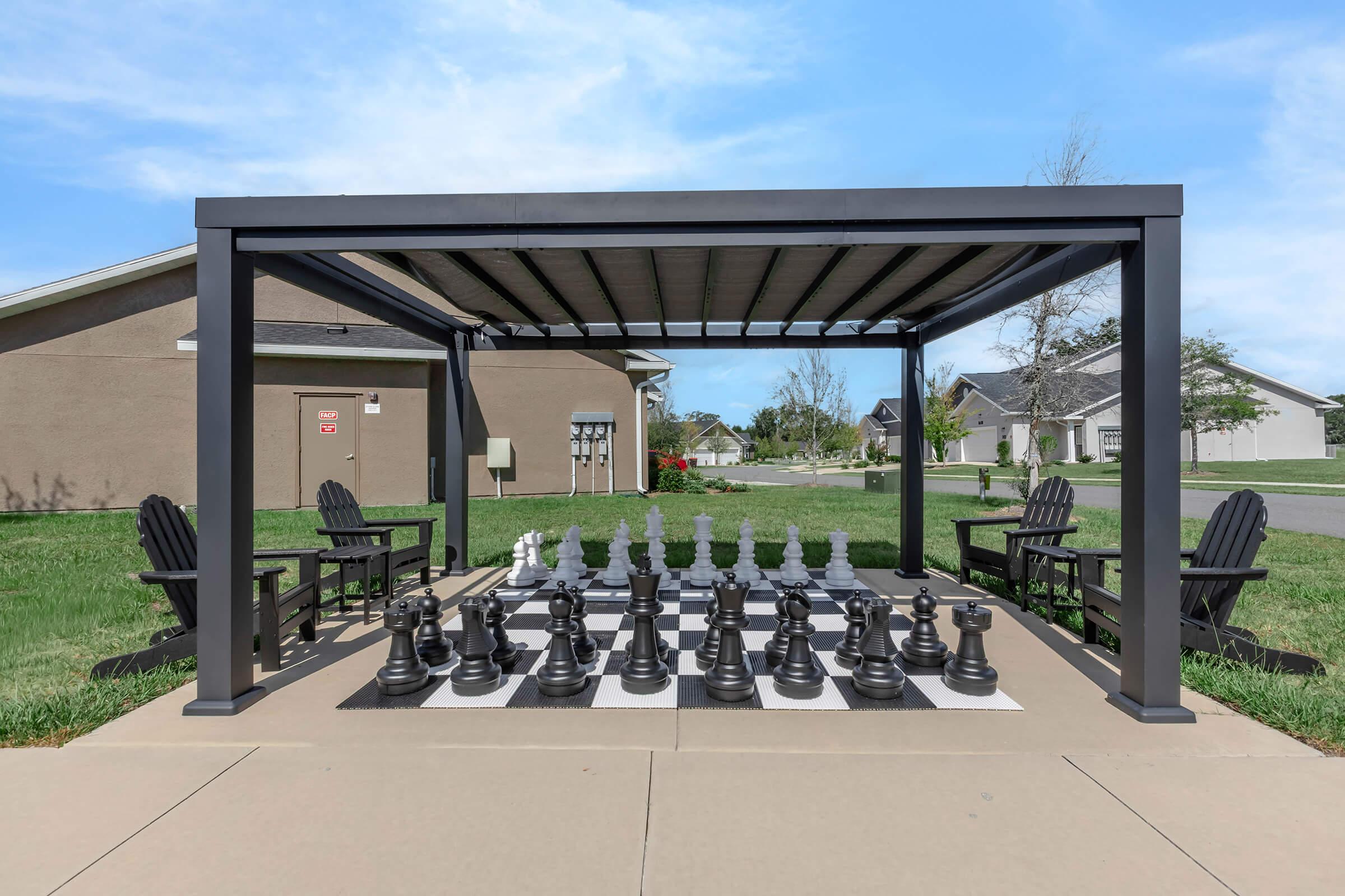 A large outdoor chess set with black and white pieces arranged on a checkered board. The setup is shaded under a pergola, with two wooden chairs nearby, surrounded by green grass and residential buildings in the background. The scene is bright and inviting.
