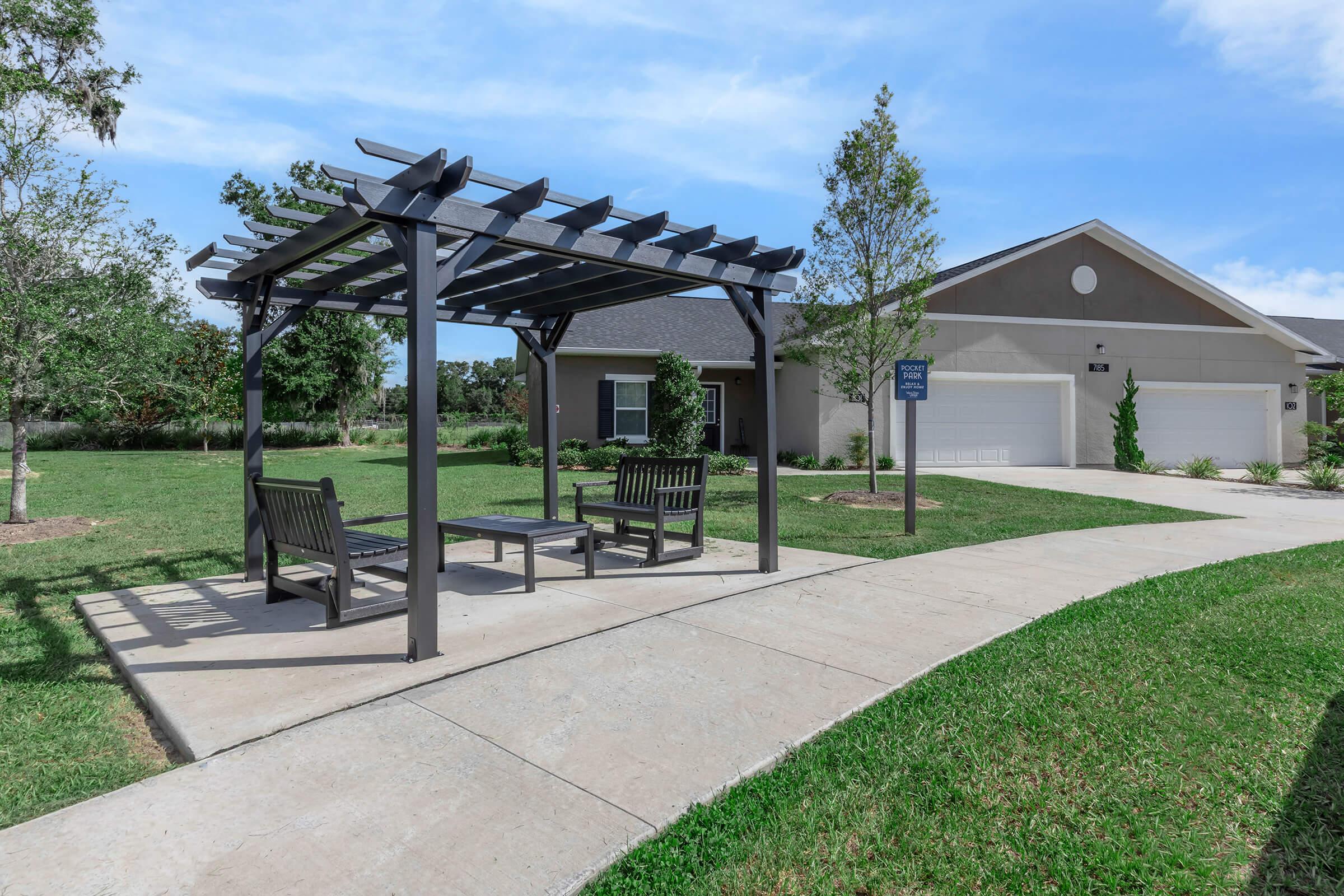 A shaded pergola with benches sits in a landscaped area beside a paved walkway. A residential building with a gray exterior and two garages is visible in the background, surrounded by green lawns and small trees under a clear blue sky.