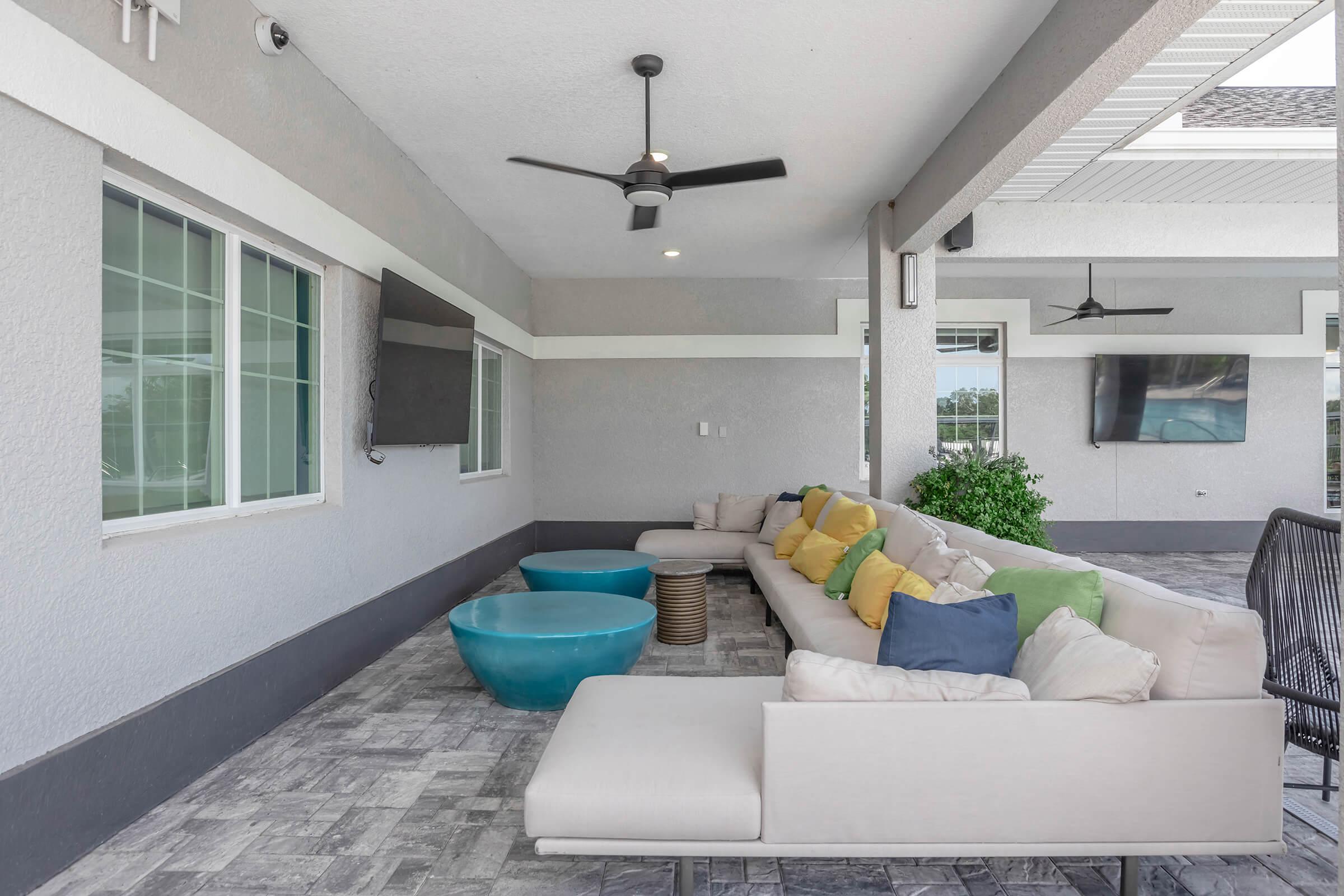 A modern outdoor lounge area featuring a spacious white sofa adorned with colorful cushions, two blue accent tables, and a ceiling fan. The walls are painted in gray tones, with large windows allowing natural light. Two wall-mounted TVs are visible in the background, creating a relaxed atmosphere.