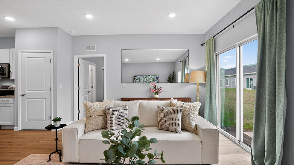 A cozy living room featuring a beige sofa with decorative pillows, a small side table with a plant, and a lamp. Large windows with green curtains overlook a grassy area. A mirror and a piece of art adorn the wall, creating a bright and inviting atmosphere. The kitchen area is visible in the background.