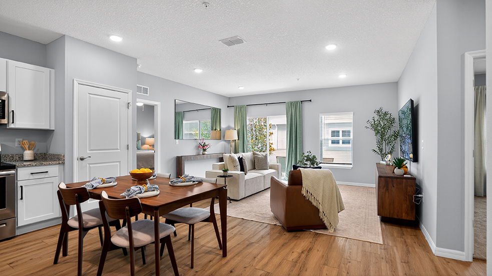 A modern living room and dining area featuring a wooden dining table set for four, a comfortable sofa, and a large TV on a wooden console. The space has light grey walls, soft lighting, and decorative plants, with green curtains framing windows that let in natural light. A cozy atmosphere with warm wooden flooring.