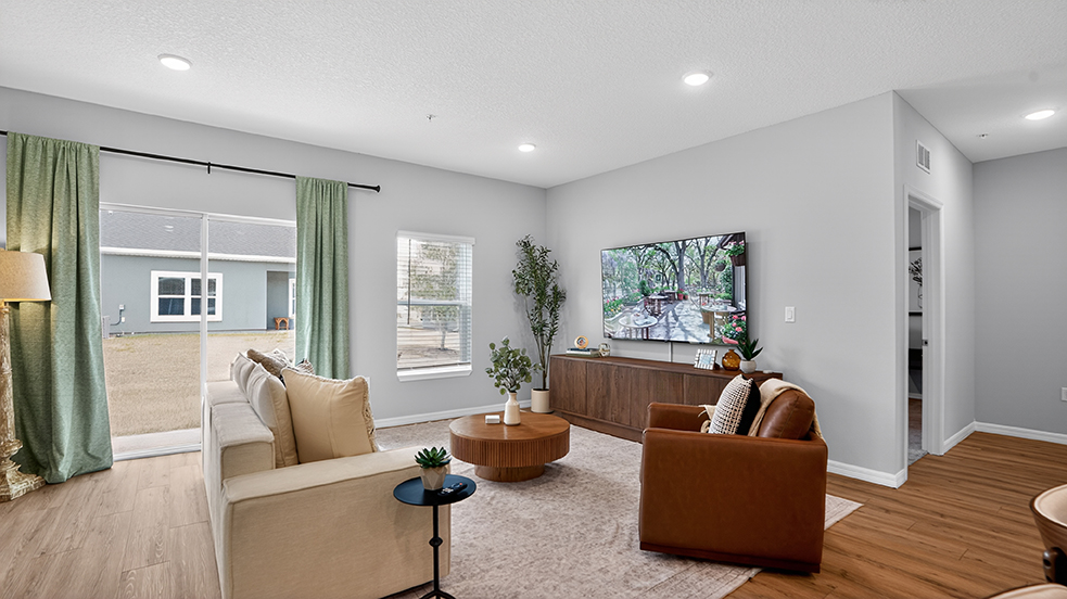 A modern living room featuring a light-colored sofa, a brown armchair, and a round wooden coffee table. Natural light fills the space through large windows with green curtains. A television mounted on the wall displays a serene outdoor scene, and the flooring is wood laminate.