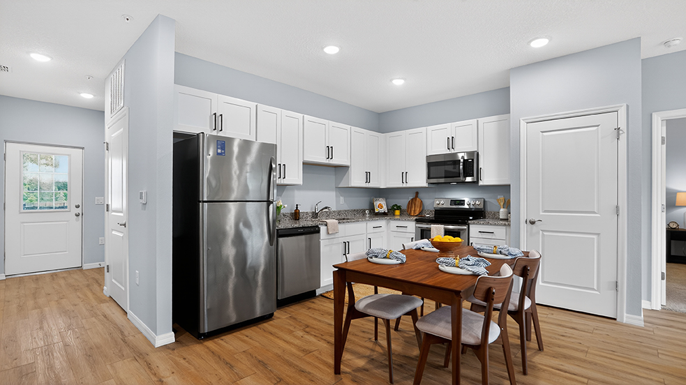 A modern kitchen featuring a stainless steel refrigerator, oven, and microwave. Bright white cabinetry contrasts with a dark countertop. A wooden dining table is set with plates and napkins, surrounded by four chairs. The space has light walls and natural wood flooring, with a door visible in the background.