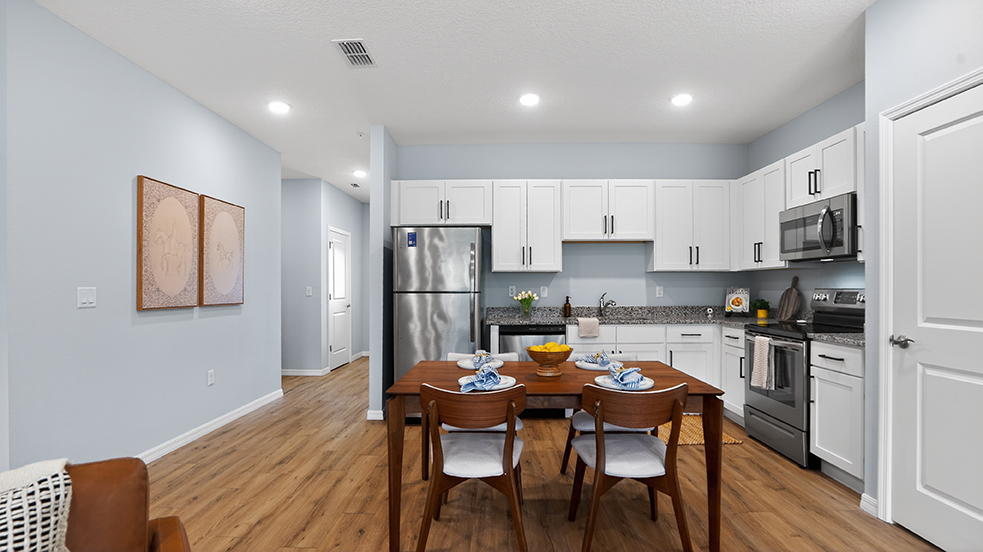 A modern, well-lit kitchen and dining area featuring a wooden dining table set for two, a sleek refrigerator, stainless steel appliances, white cabinetry, and light blue walls. A cozy chair is in the foreground, and decorative art hangs on the wall. The space is inviting and stylish, with natural light enhancing the ambiance.