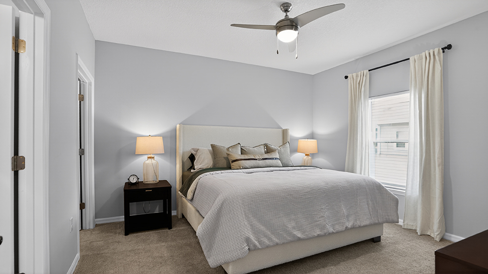 A modern bedroom featuring a large upholstered bed with decorative pillows, two bedside lamps, and a nightstand. There are light gray walls, a ceiling fan, and light-colored curtains. The room has a warm, inviting atmosphere with soft carpeting and well-placed windows allowing natural light.