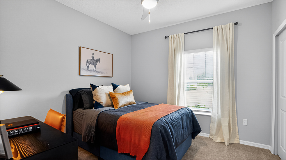 A cozy bedroom featuring a bed with dark blue and orange bedding, decorative pillows, and a large painting of a horse on the wall. Natural light comes through a window with sheer curtains, and there's a bedside table with a lamp and a magazine. The walls are painted light gray.