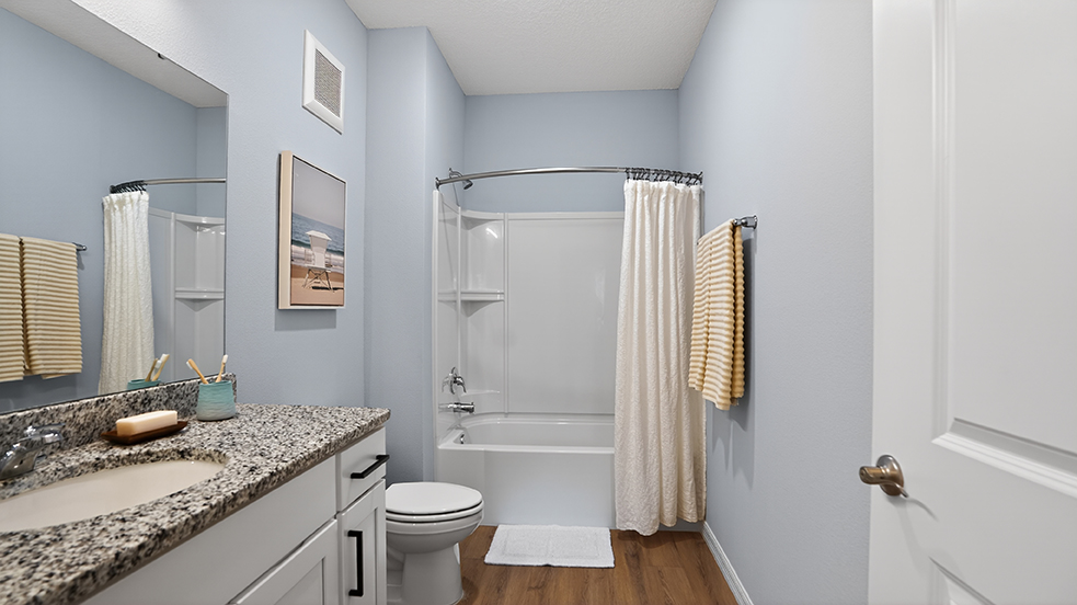 A modern bathroom featuring a light blue wall, a white bathtub with a shower curtain, a granite countertop with a sink, and a round mirror. There are beige towels hanging, a beach-themed picture on the wall, and wooden flooring, creating a bright and inviting atmosphere.