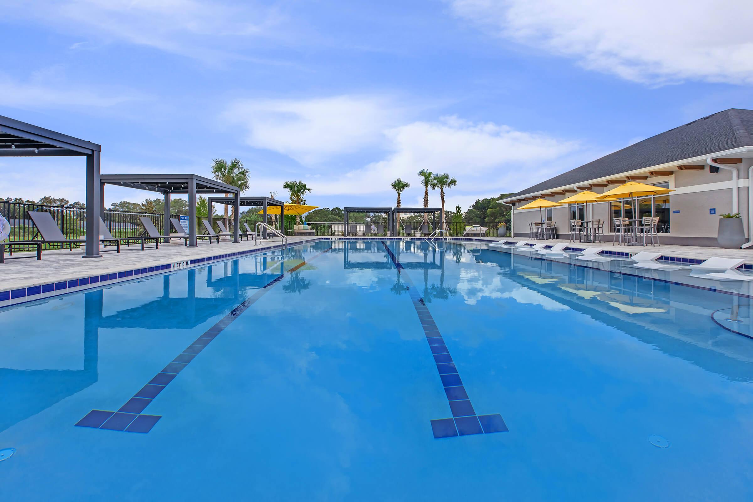 A clear swimming pool with lanes marked by dark blue tiles, surrounded by lounge chairs and shaded areas. Palm trees line the background, and a modern building with yellow awnings is visible on the right. The sky is bright with a few clouds, creating a relaxing outdoor atmosphere.