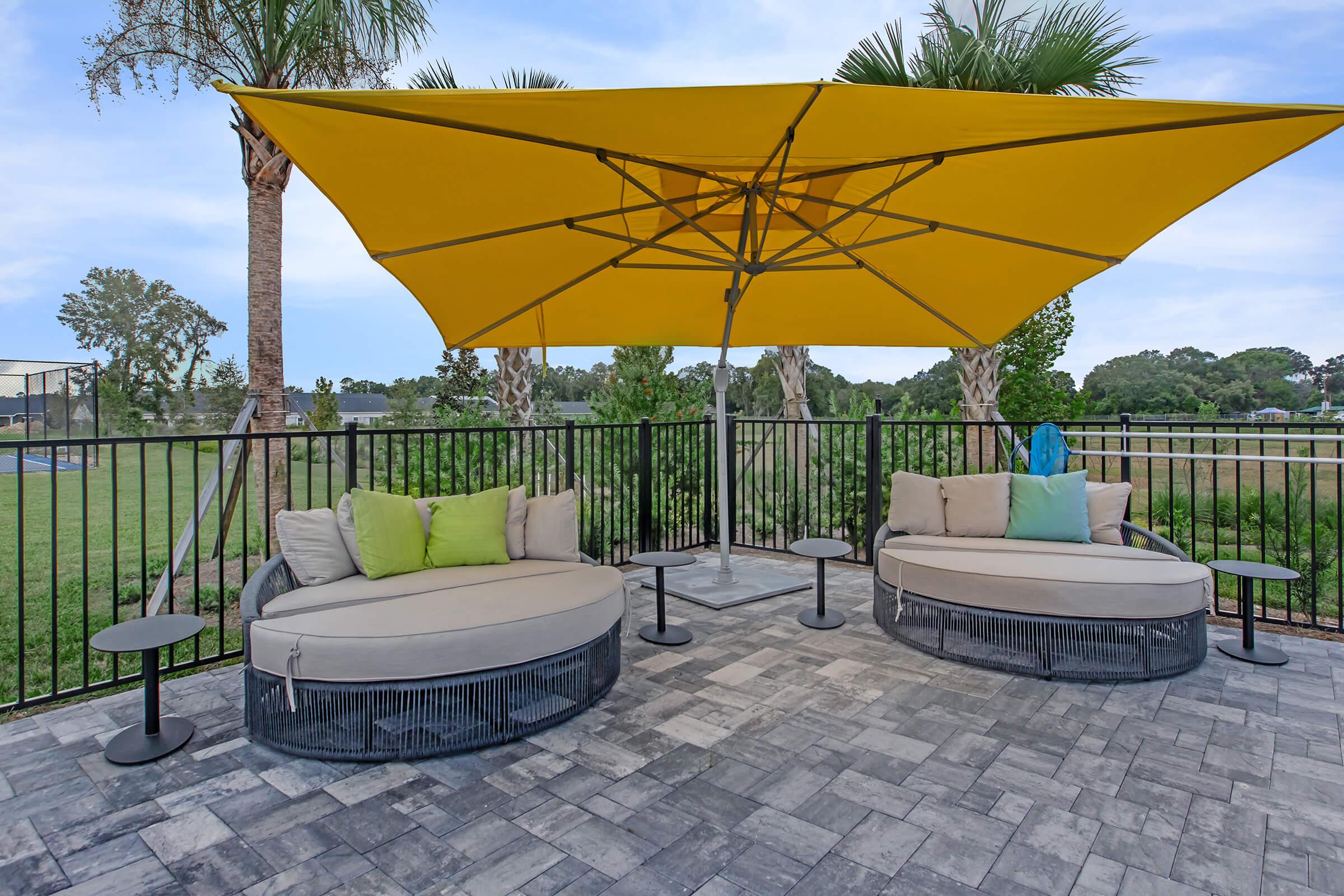 Outdoor seating area featuring two circular lounge chairs with light-colored cushions and vibrant green throw pillows. A large yellow umbrella provides shade over the seating. The space is surrounded by a black fence, palm trees, and a green landscape in the background, creating a relaxing atmosphere.
