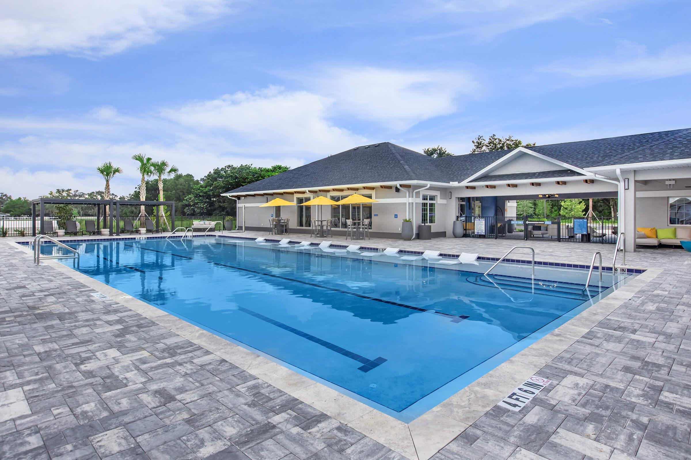 A clear blue swimming pool surrounded by a spacious patio area. Yellow umbrellas provide shade near the poolside. A modern building with a dark roof is visible in the background, featuring large windows and a welcoming entrance. Lush greenery can be seen beyond the pool area under a bright, blue sky.