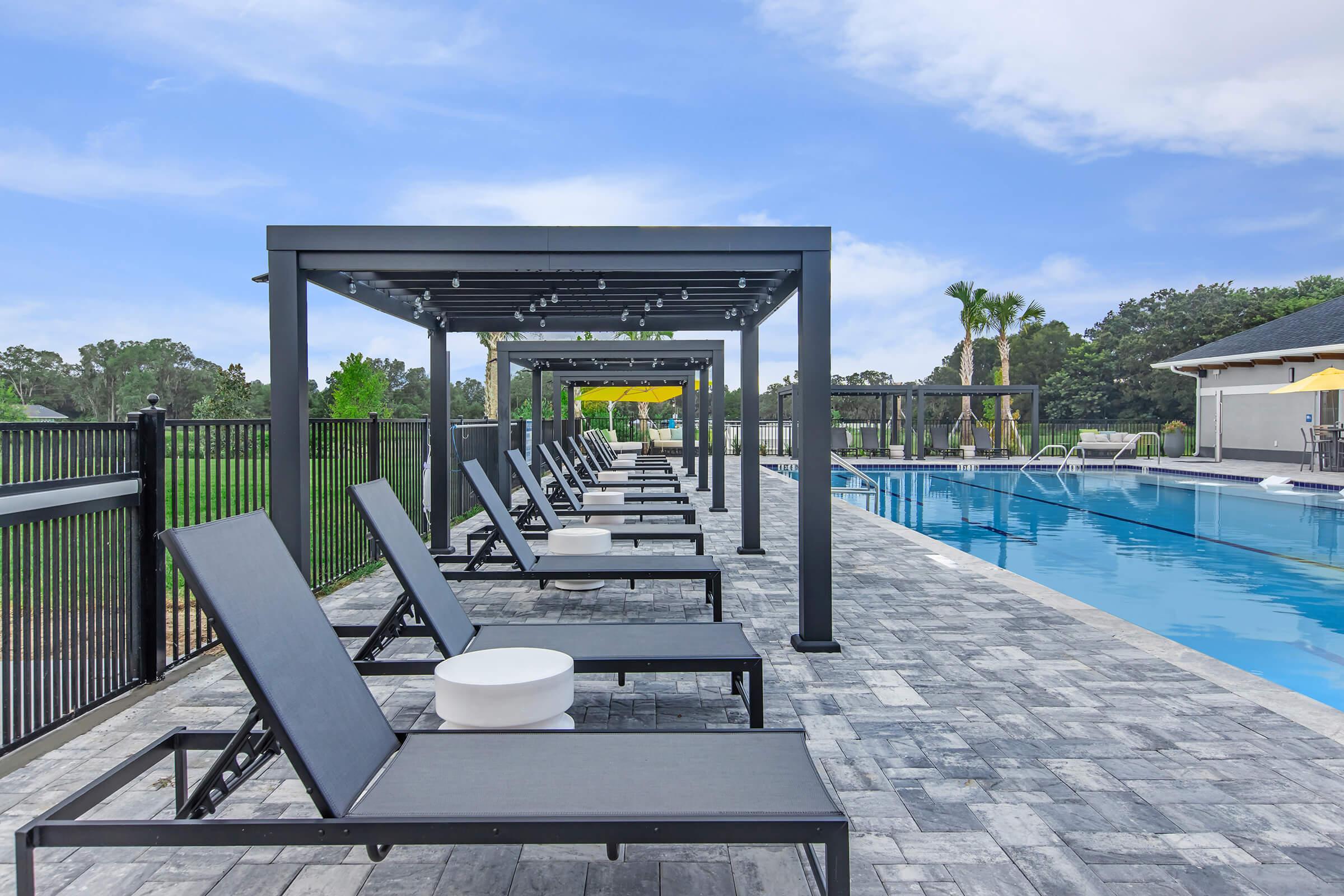 A serene pool area featuring a row of black lounge chairs under shaded canopies. The pool, lined with vibrant green landscaping, reflects the clear blue sky. Yellow umbrellas provide additional shade, creating a relaxing atmosphere for leisure and enjoyment.